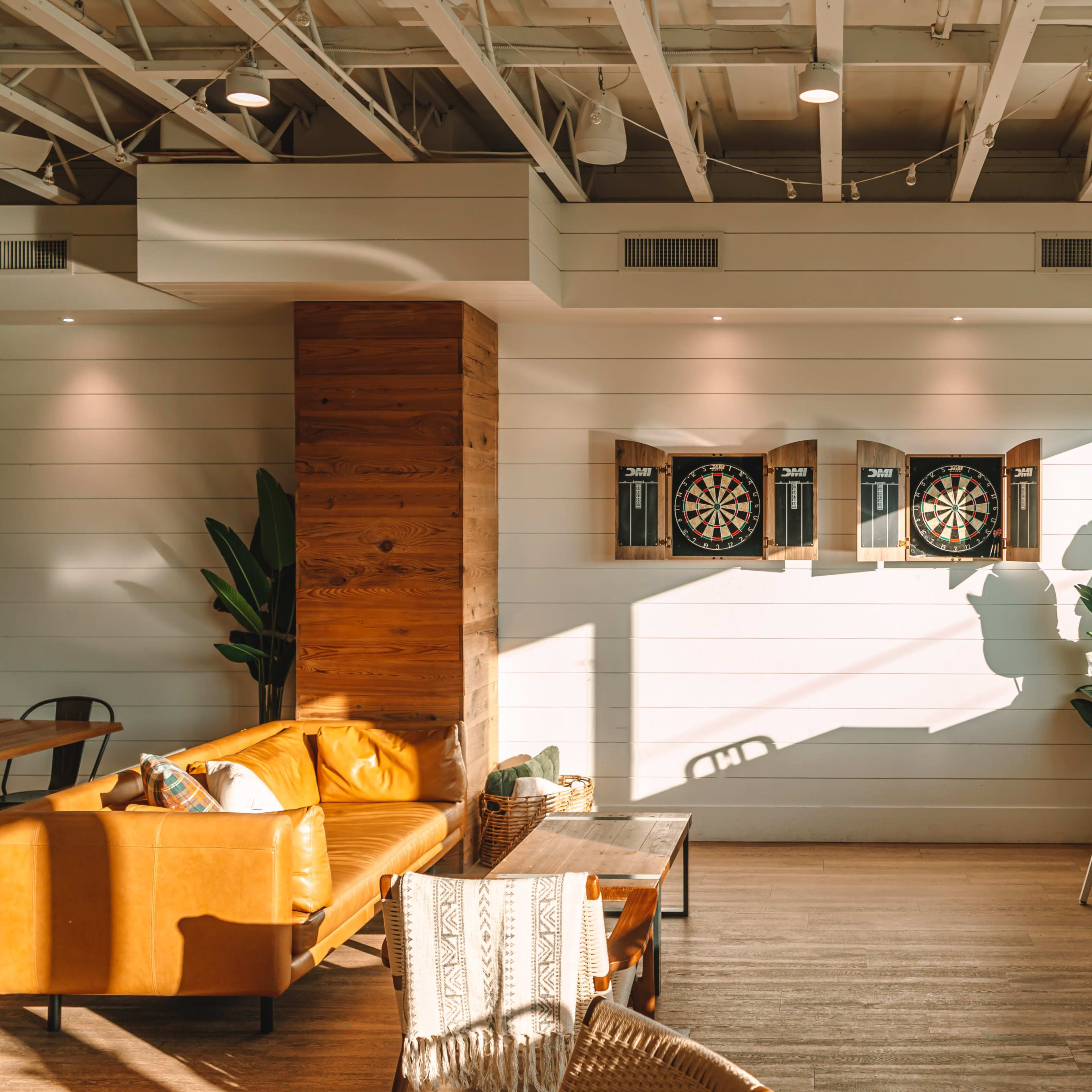 A cozy interior features a seating area with a leather couch, dart boards on the wall, and large windows allowing natural light to illuminate the space.