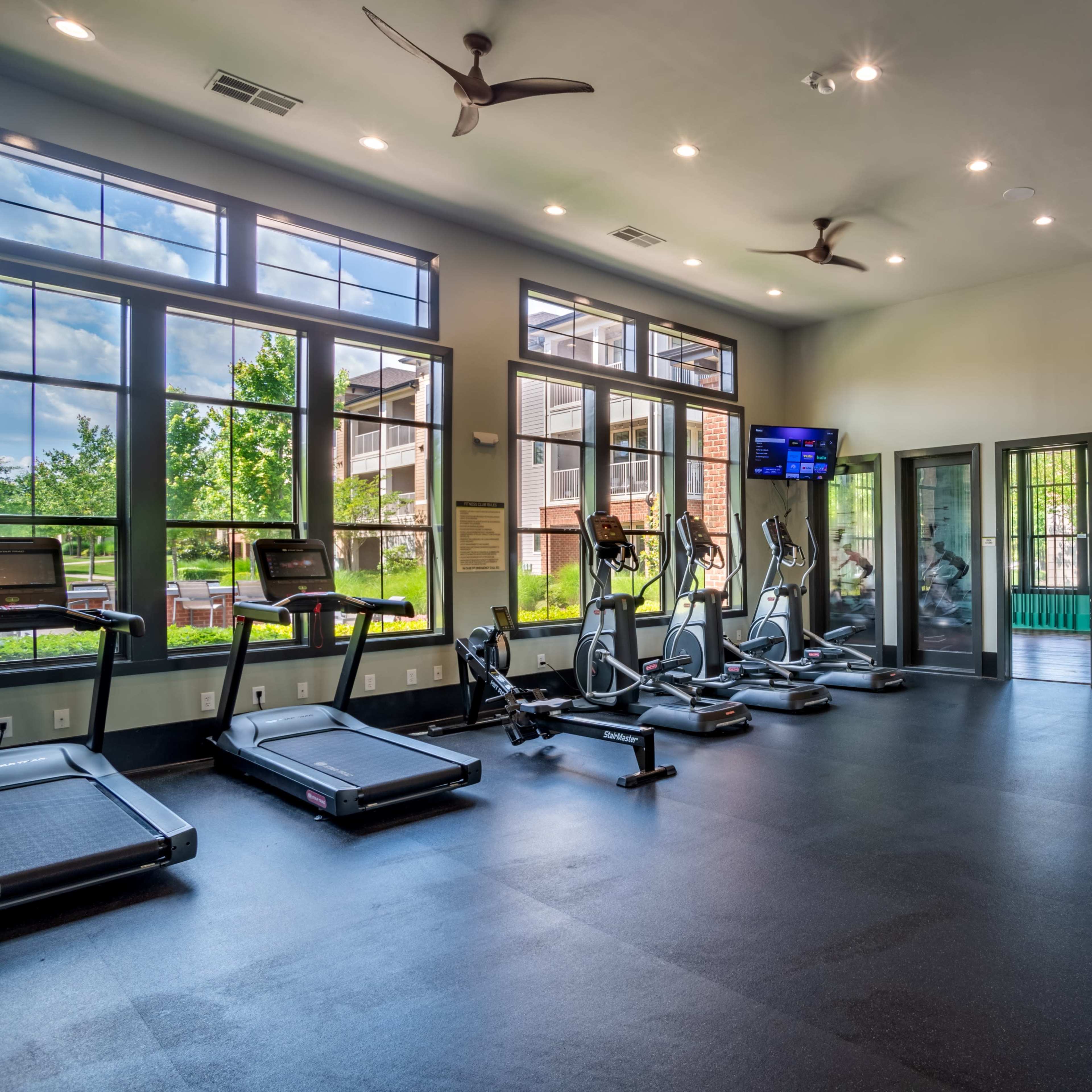 A modern gym with exercise equipment, including treadmills and elliptical machines, in a well-lit space featuring large windows.