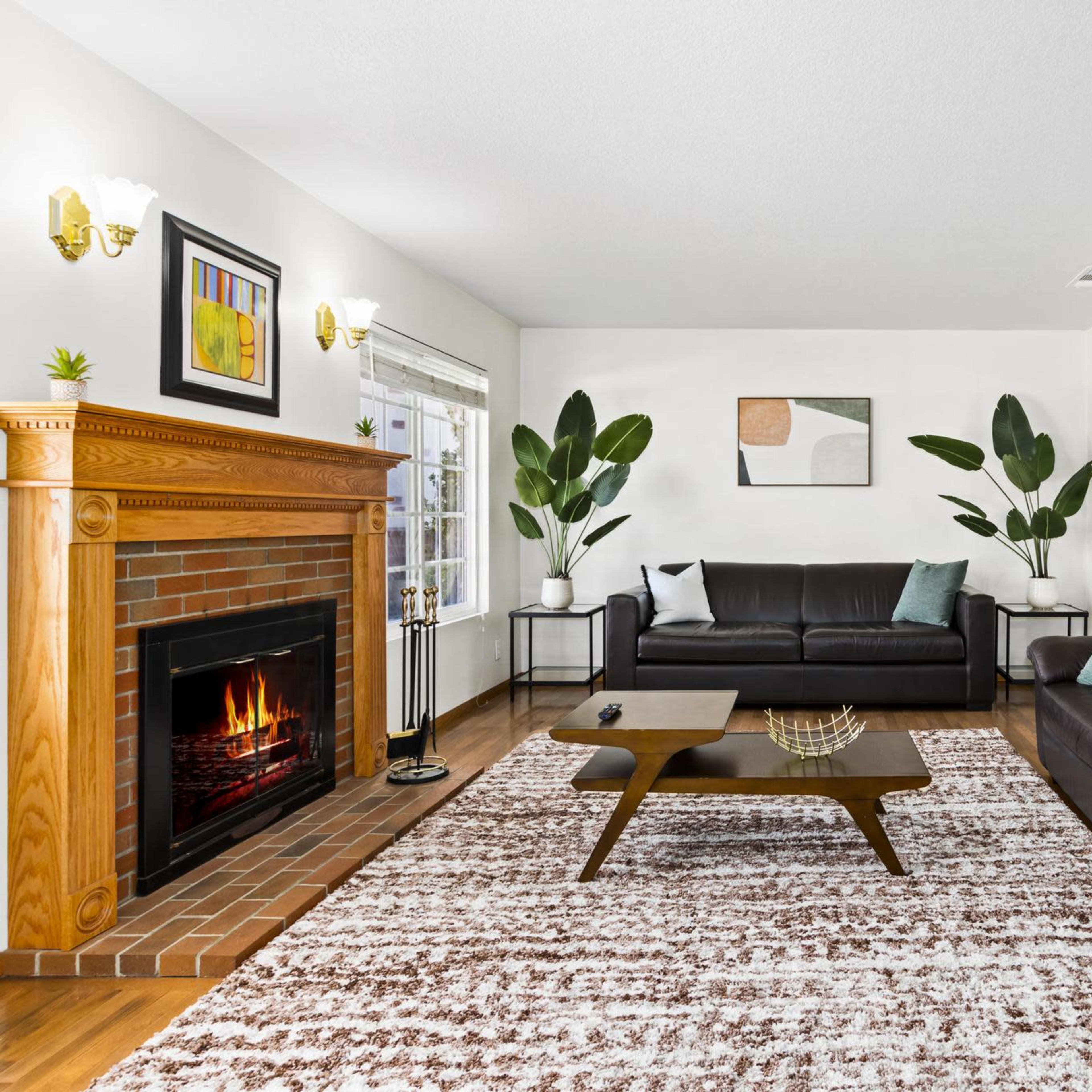 A living room with a brick fireplace, two leather sofas, a coffee table, and plants beside large windows.