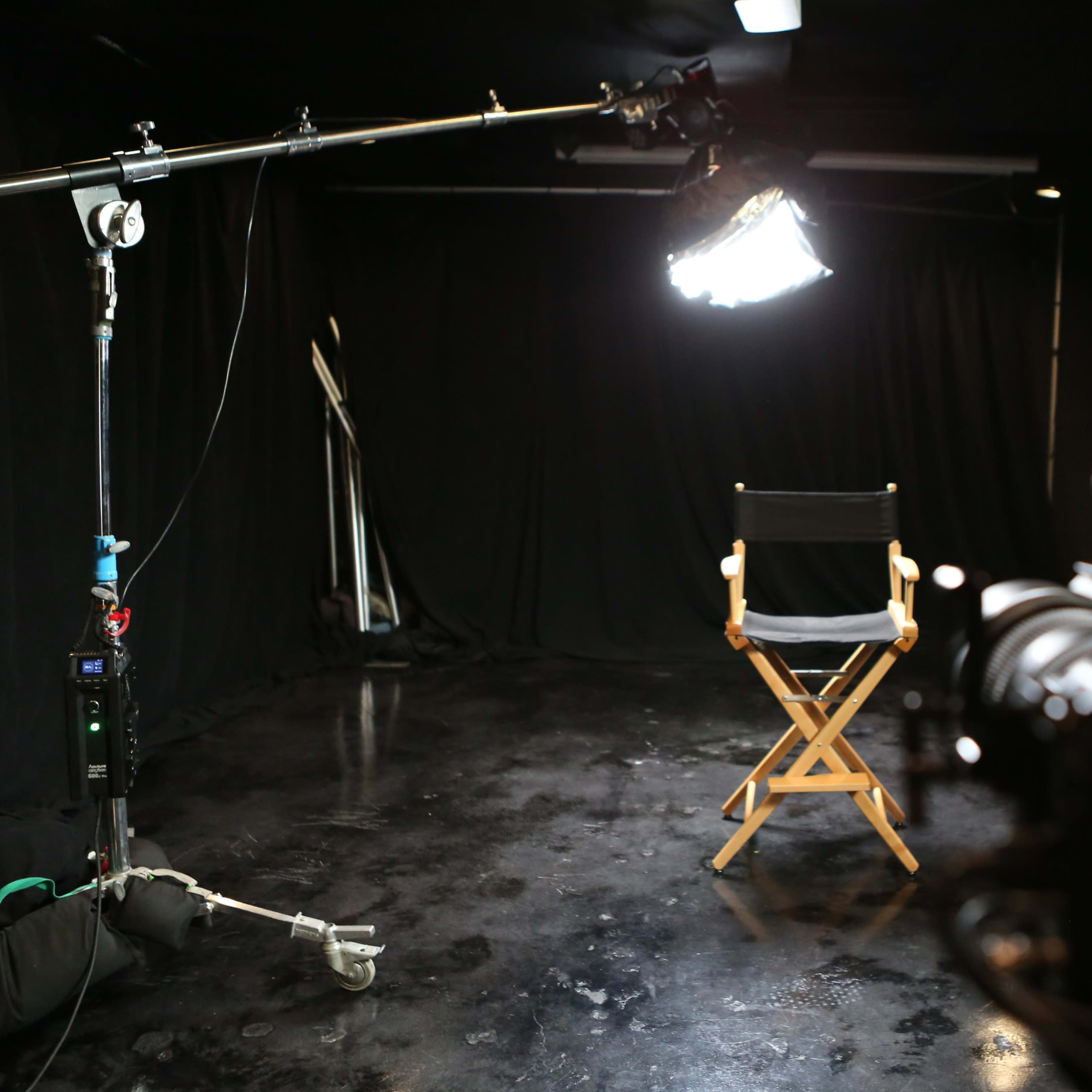 A film studio setup with a wooden director's chair facing a camera, illuminated by a large light source against a backdrop of black curtains.