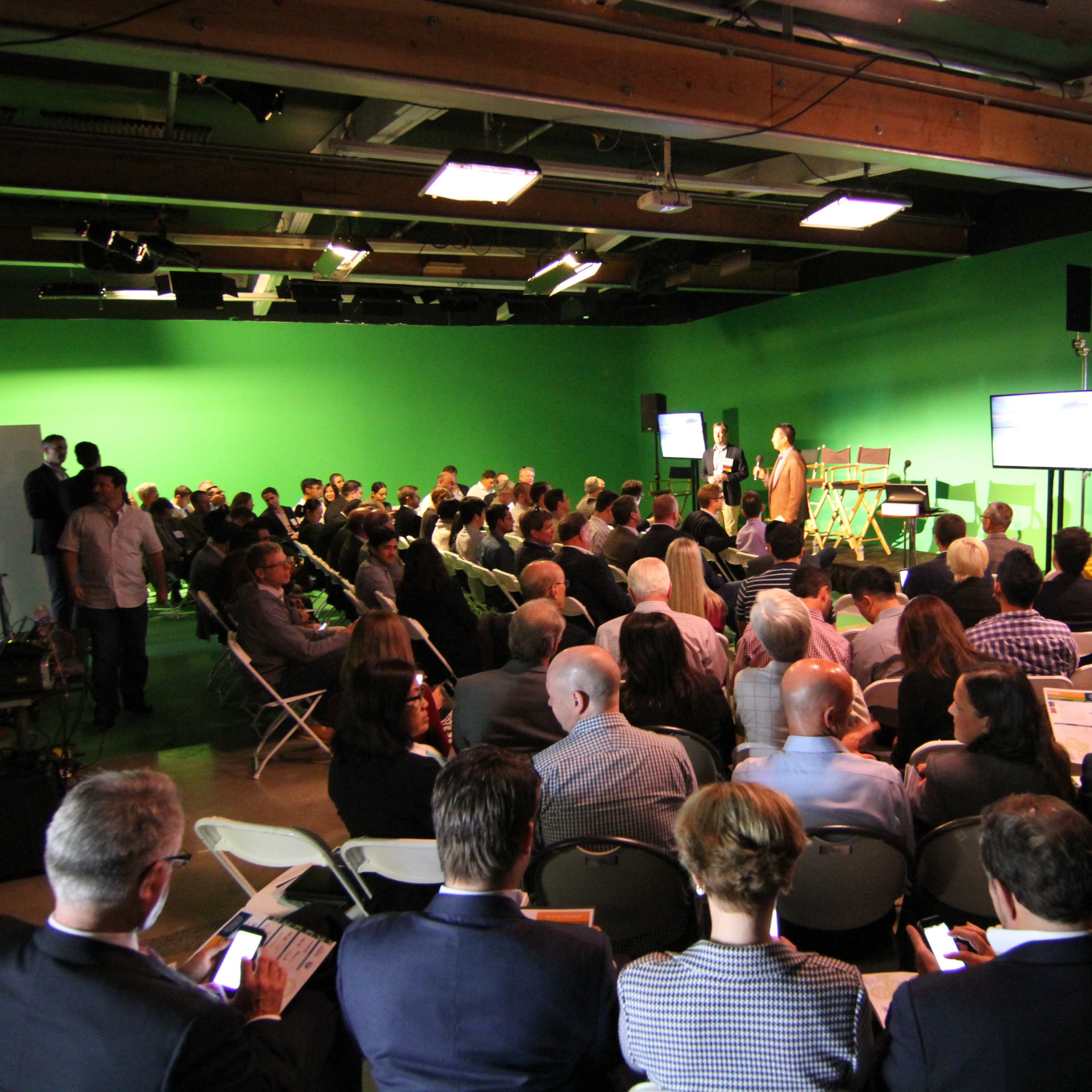 A large audience attends a presentation in a studio with a green screen backdrop, while cameras and screens are set up for the event.