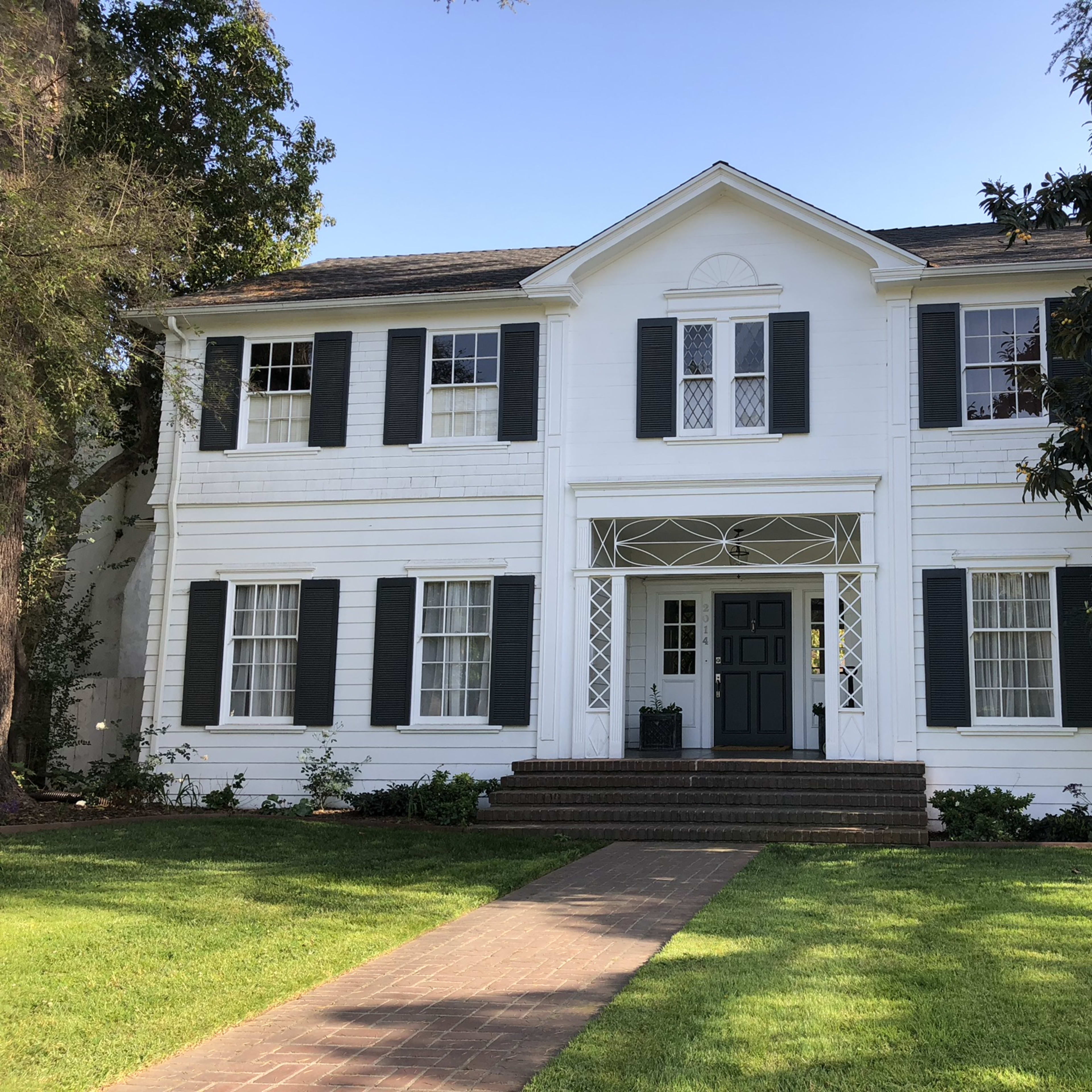 A two-story white house with green shutters and a front porch features a centered door and symmetrical window arrangement.