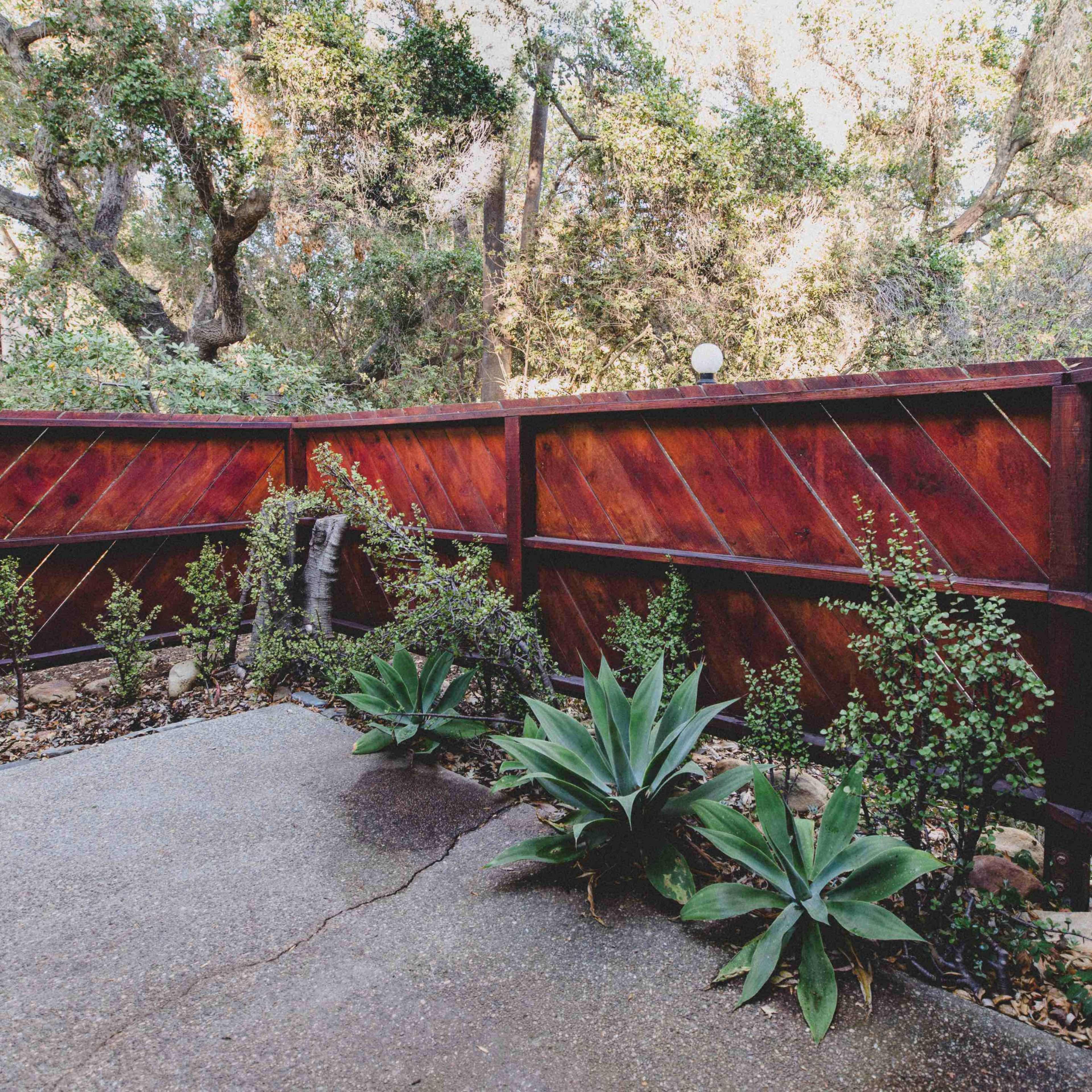 A small outdoor space enclosed by a wooden fence, featuring a concrete floor, potted plants, and a wall fountain.