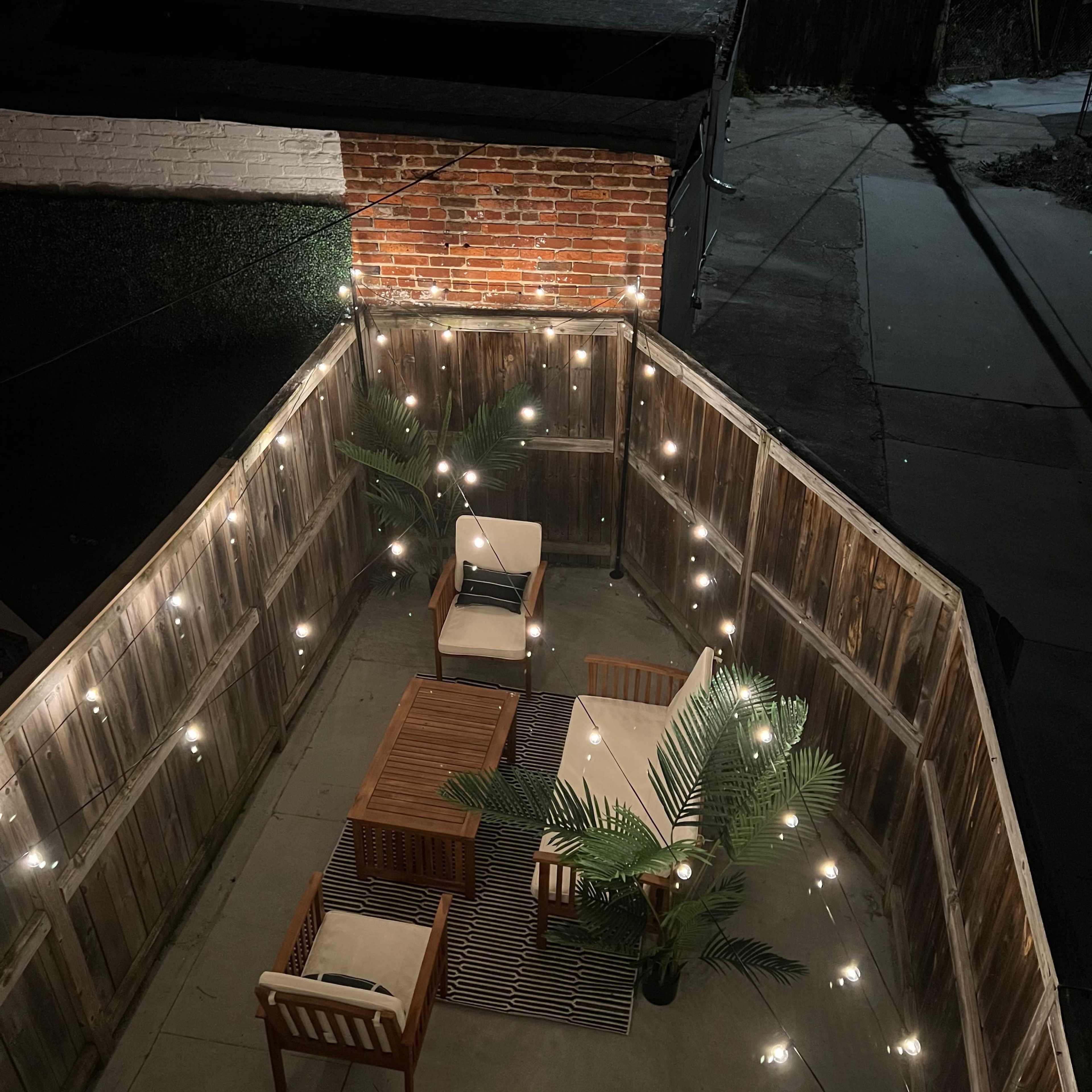 A small outdoor patio area illuminated by string lights, featuring wooden furniture and potted plants.