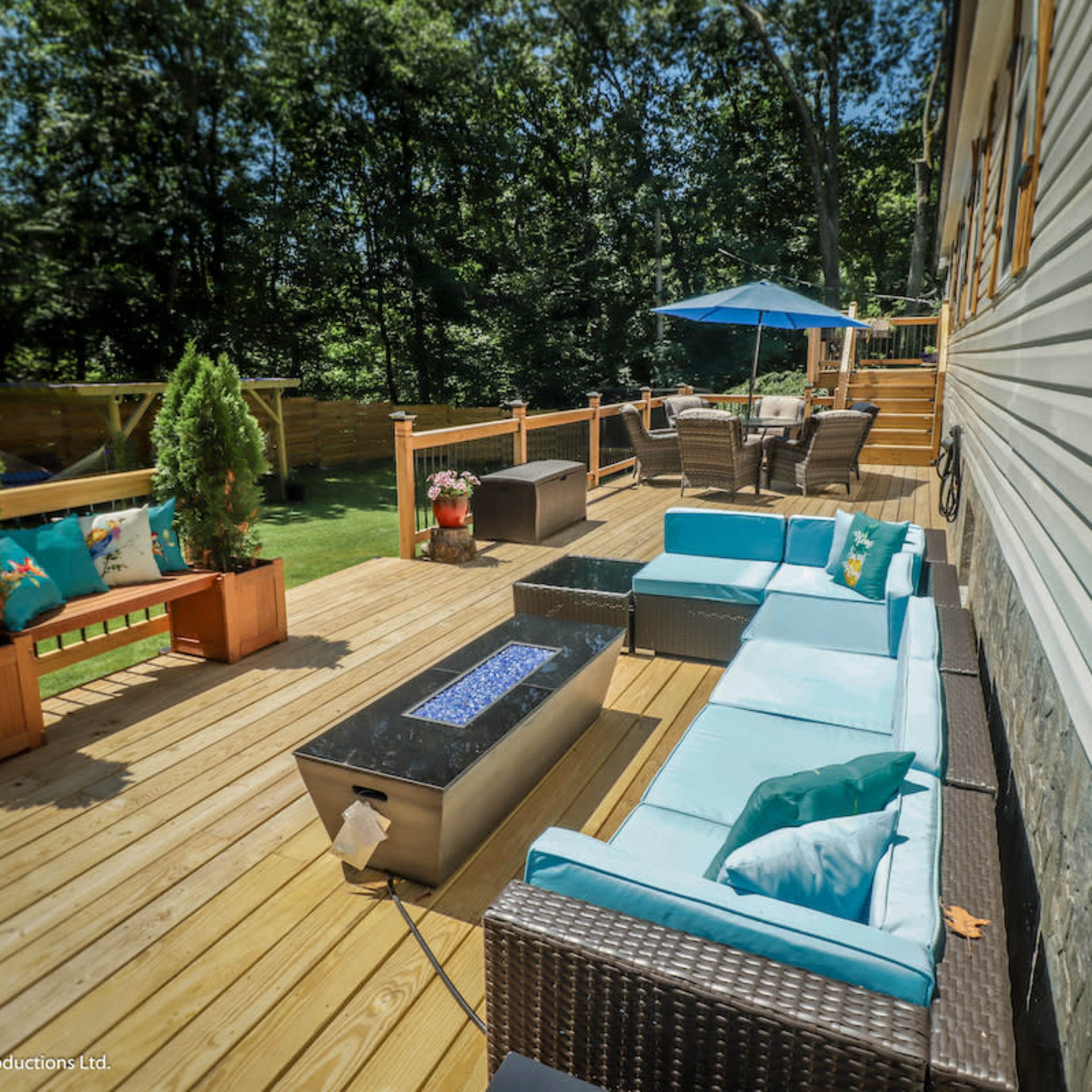 A spacious wooden deck features comfortable seating, a fire table, and an umbrella over a dining area surrounded by greenery.