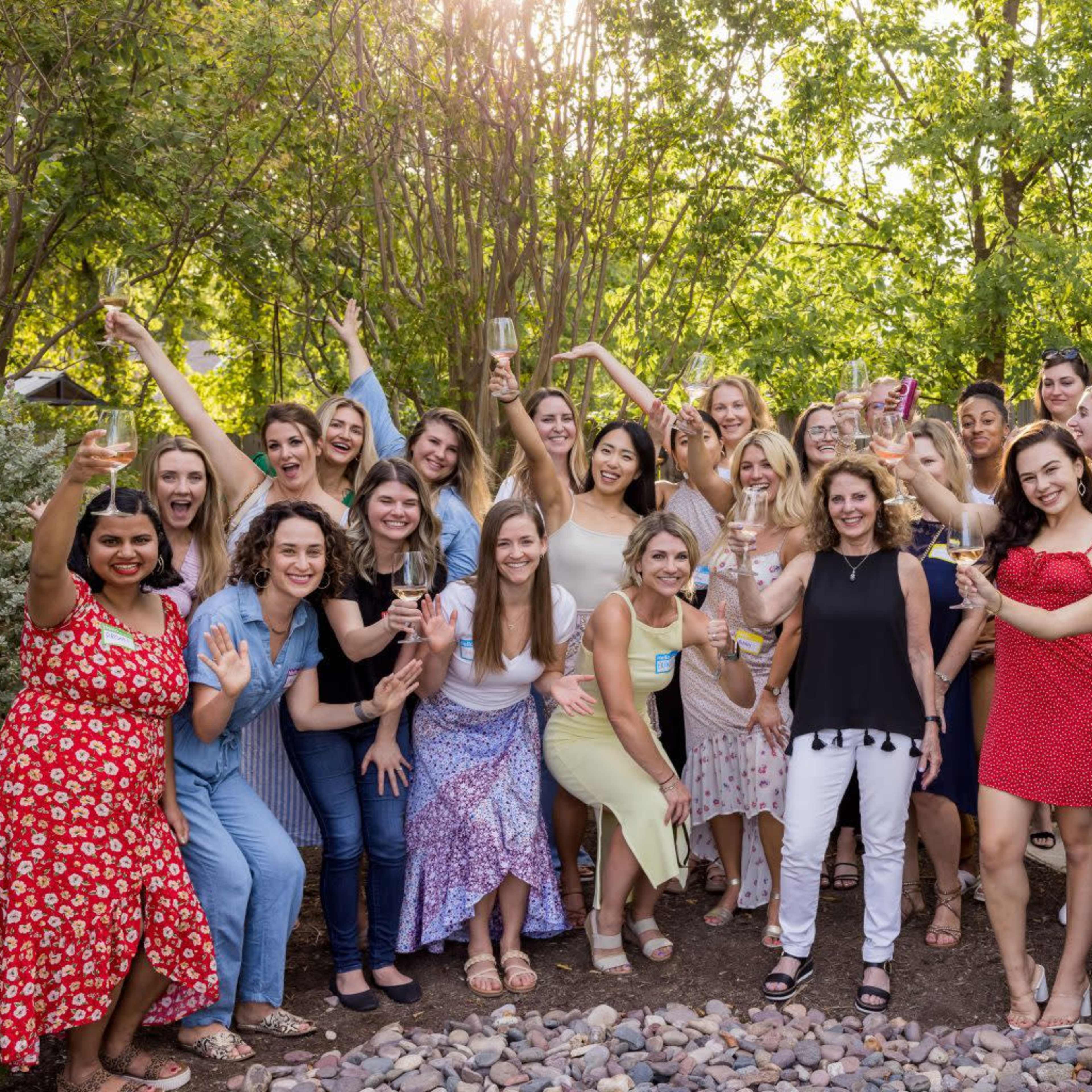 A large group of women is gathered in a backyard, smiling and posing for the camera, while holding drinks.