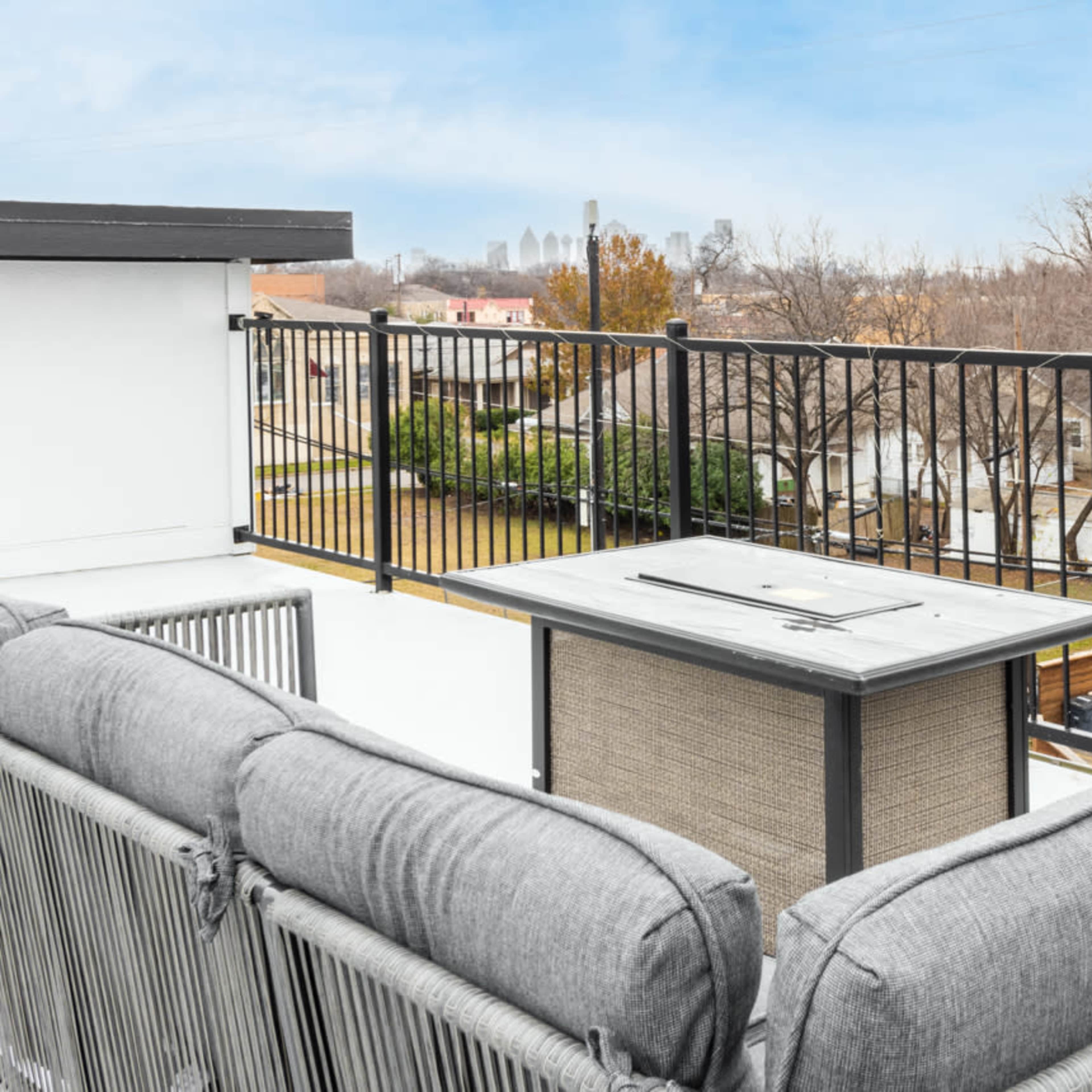 A rooftop patio with a seating area and a fire pit, overlooking a cityscape and residential backyards.