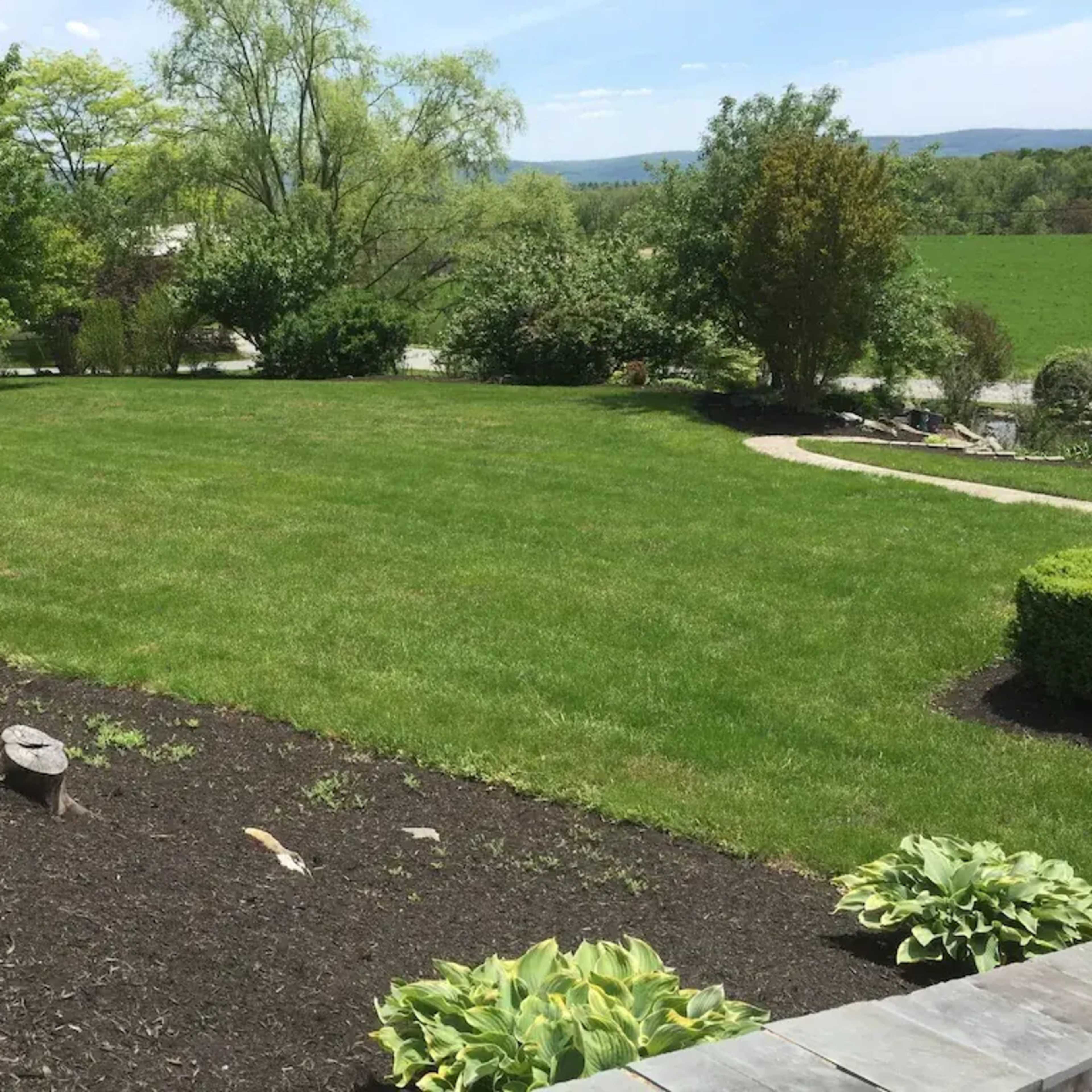 A grassy lawn is bordered by landscaped flower beds and trees, with rolling hills visible in the background.