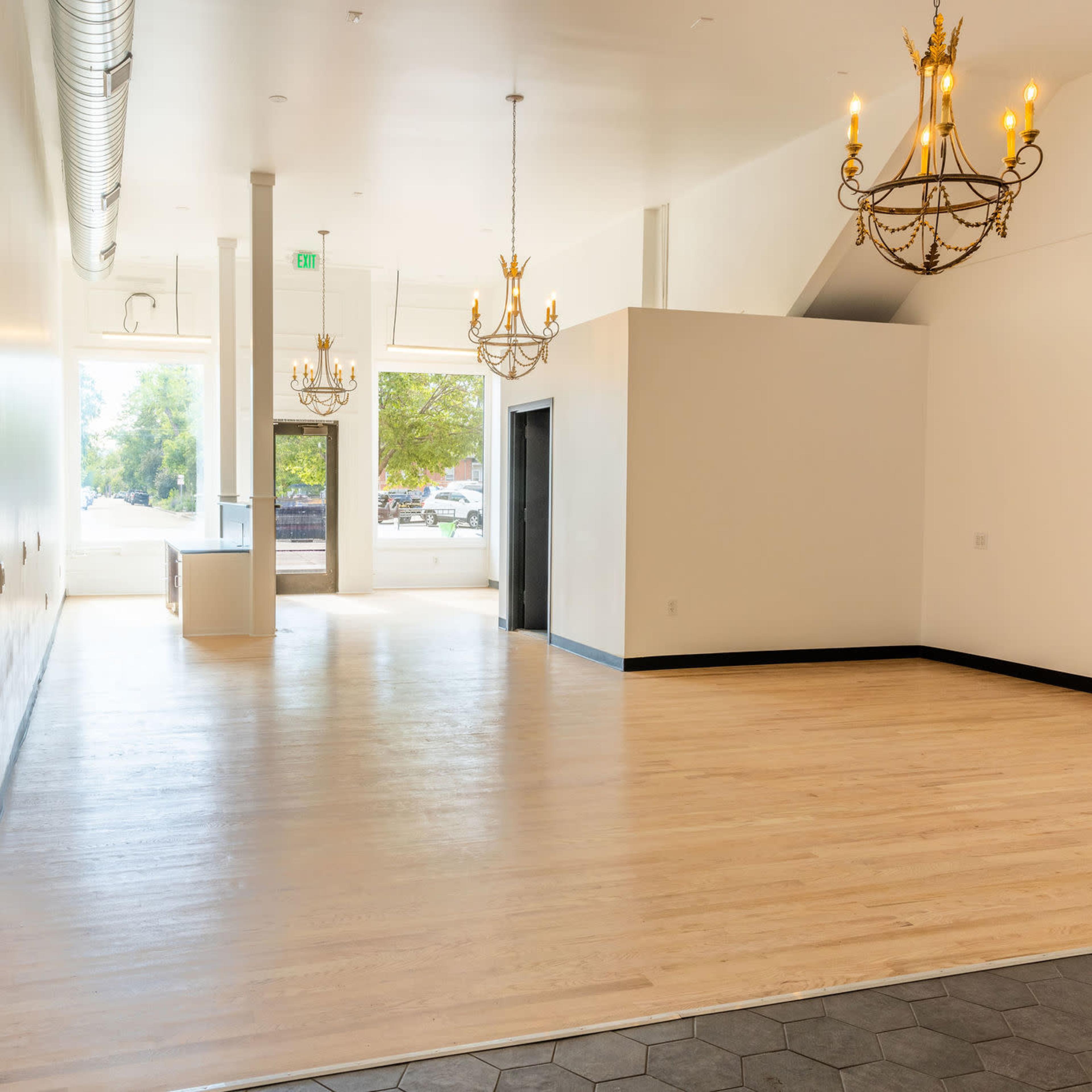 A spacious, empty room with wooden floors, large windows, and two chandeliers hanging from the ceiling.