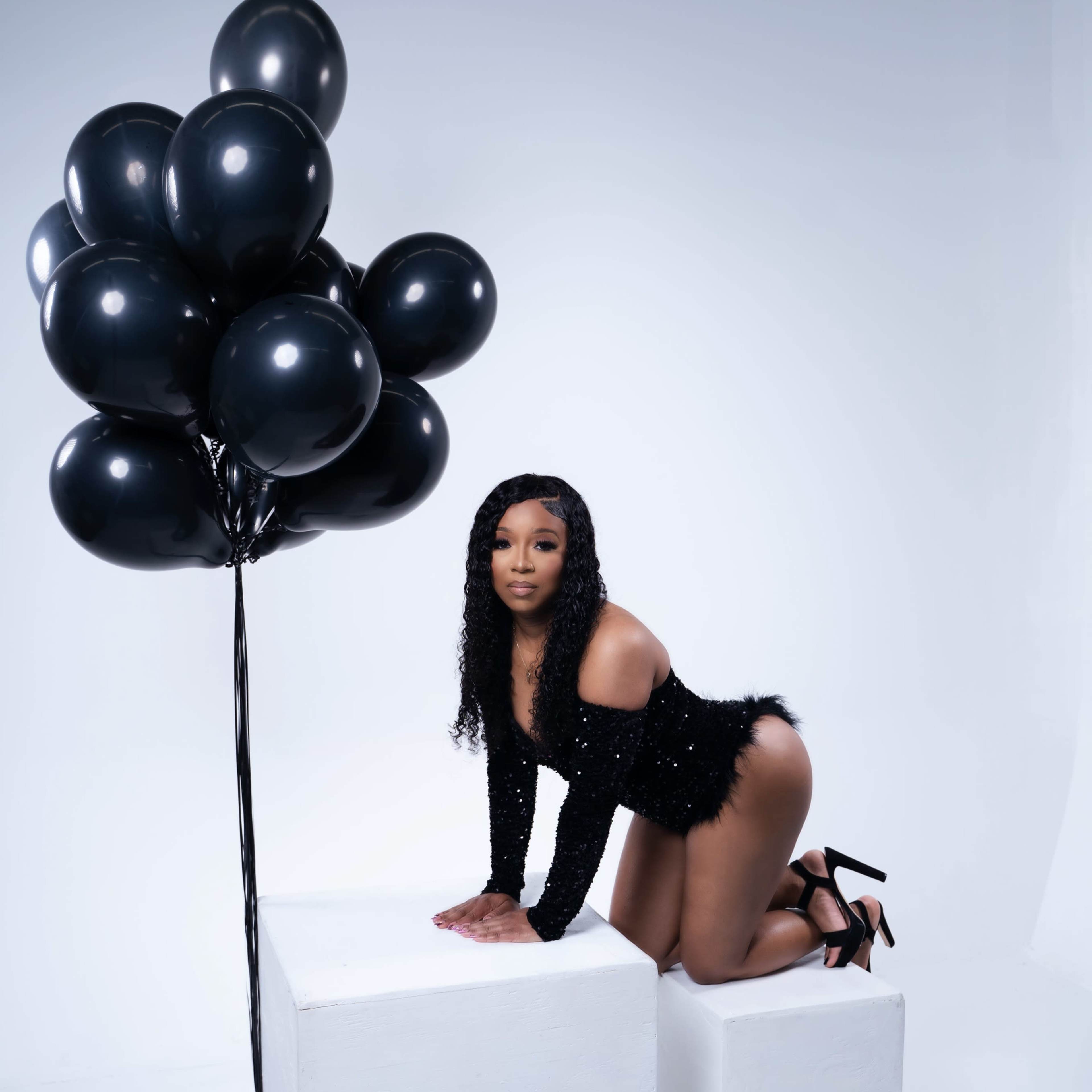 A woman in a black outfit kneels on two white cubes beside a cluster of black balloons.