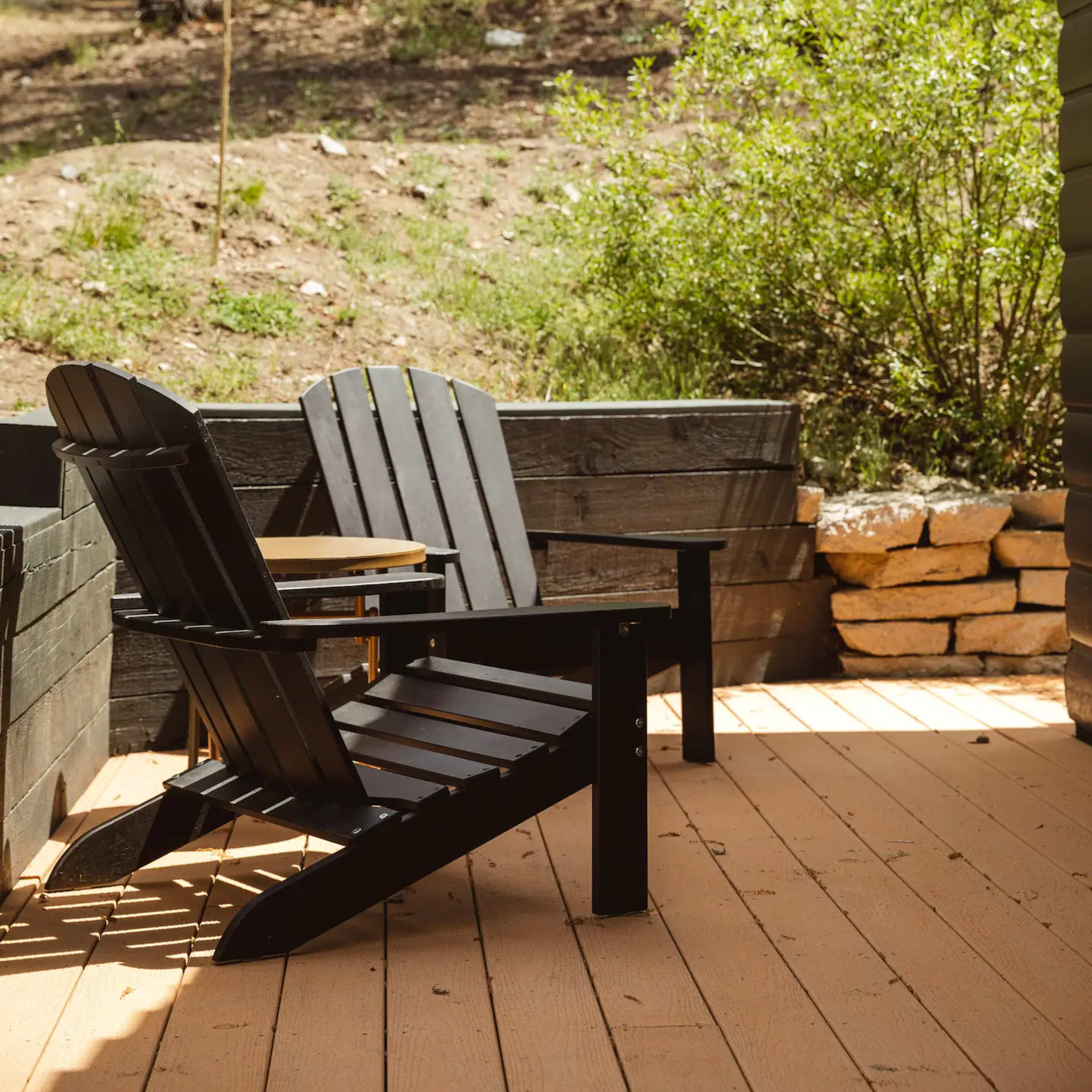 Two black Adirondack chairs sit on a wooden deck near a stone wall, surrounded by greenery and trees in the background.