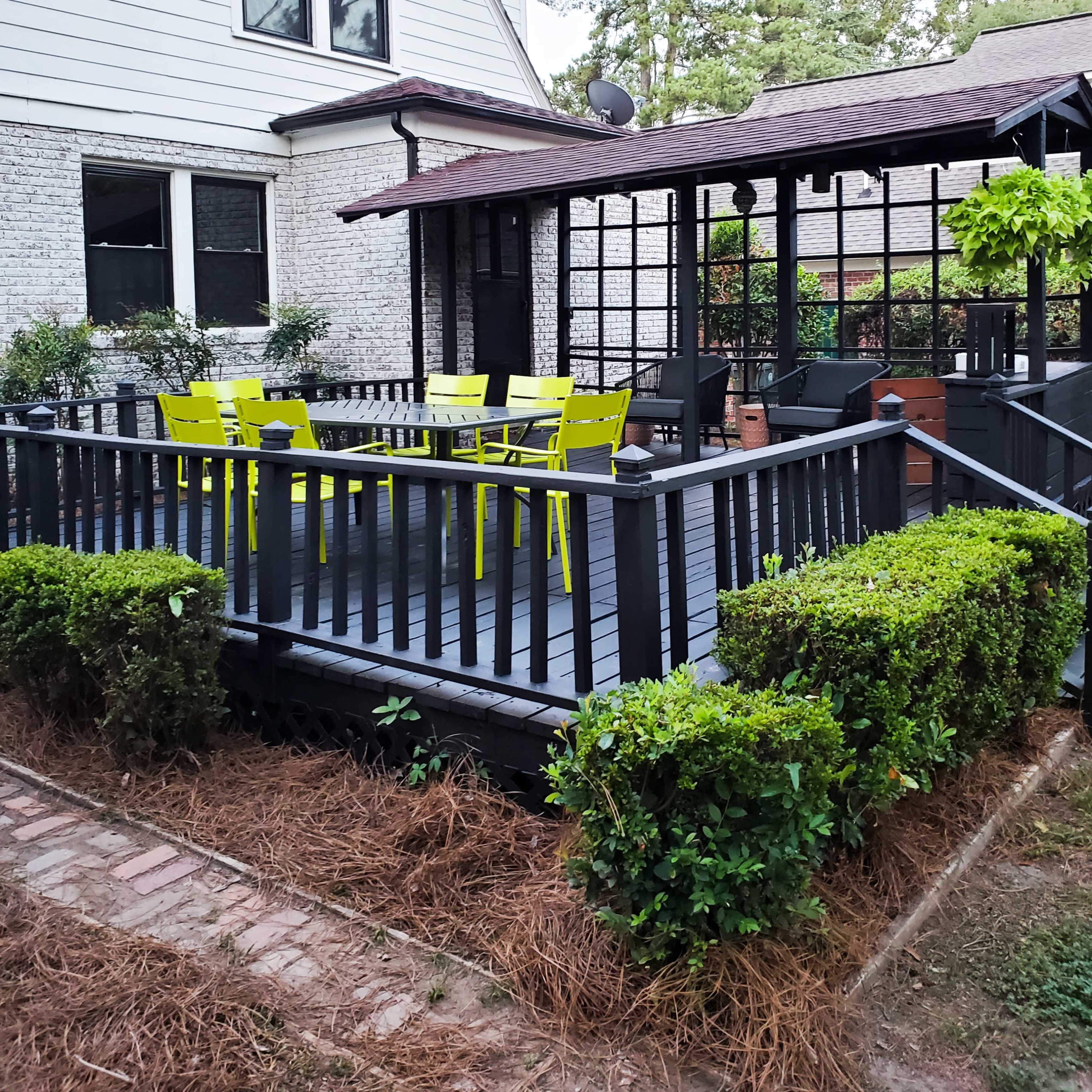 A black wooden deck with bright yellow chairs and a gazebo, surrounded by neatly trimmed bushes and a pathway made of brick.