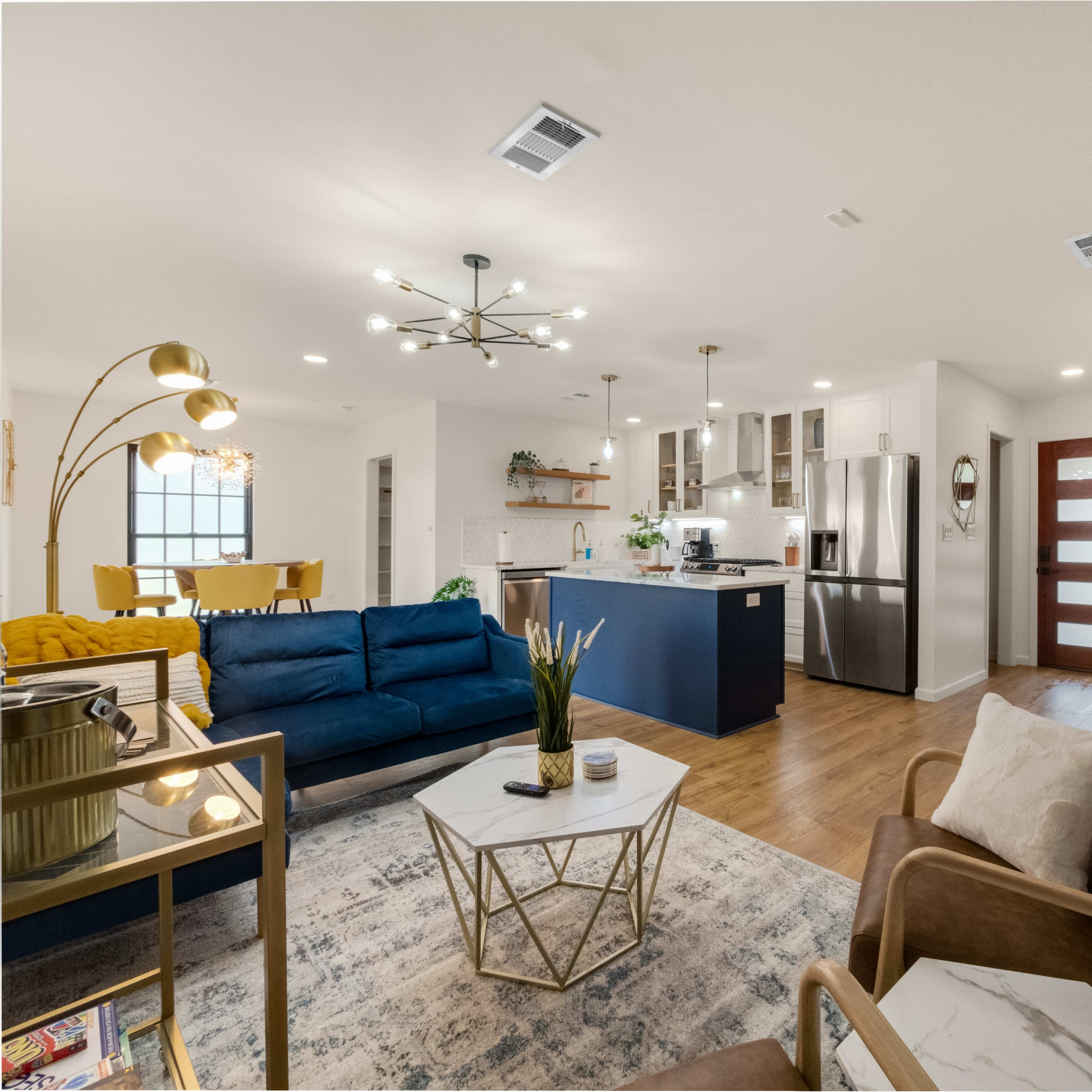 A modern living room with a blue sofa, a coffee table, and an open kitchen area featuring stainless steel appliances and a dining table in the background.