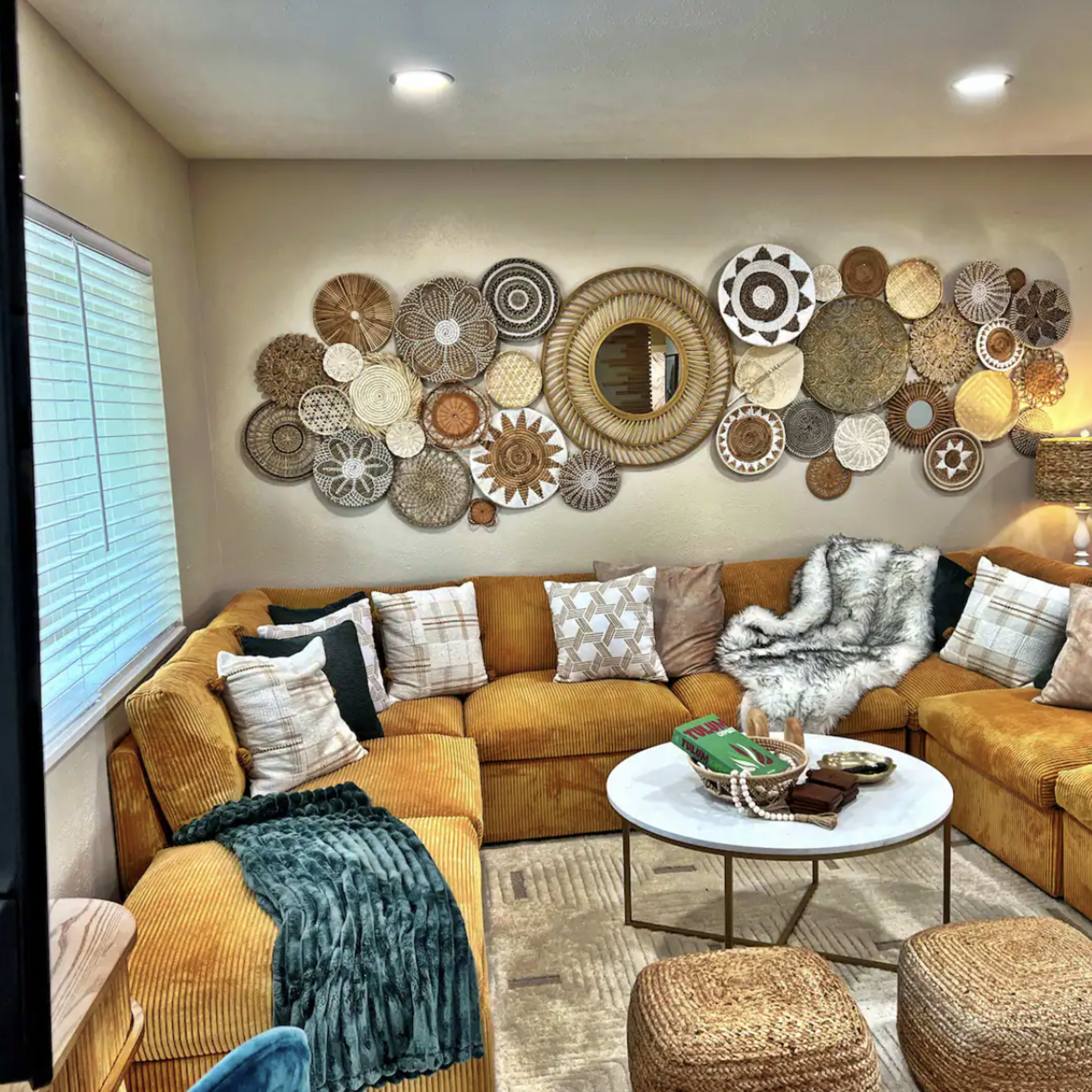 A cozy living room featuring a large, L-shaped, mustard-colored couch adorned with various patterned pillows, a round coffee table, and decorative wall art made of woven materials.