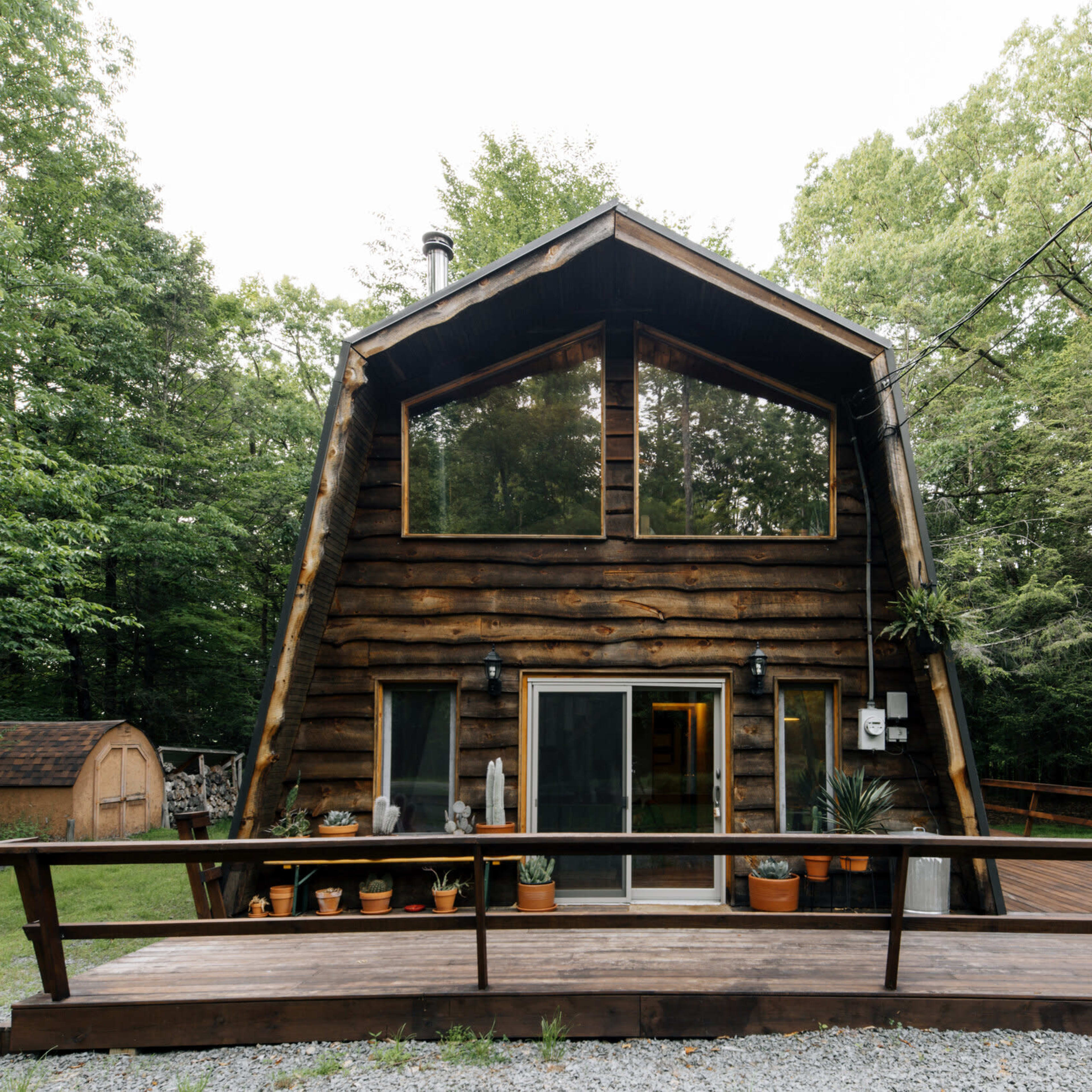 A wooden cabin with a peaked roof is set in a forested area, featuring a front deck and large windows.