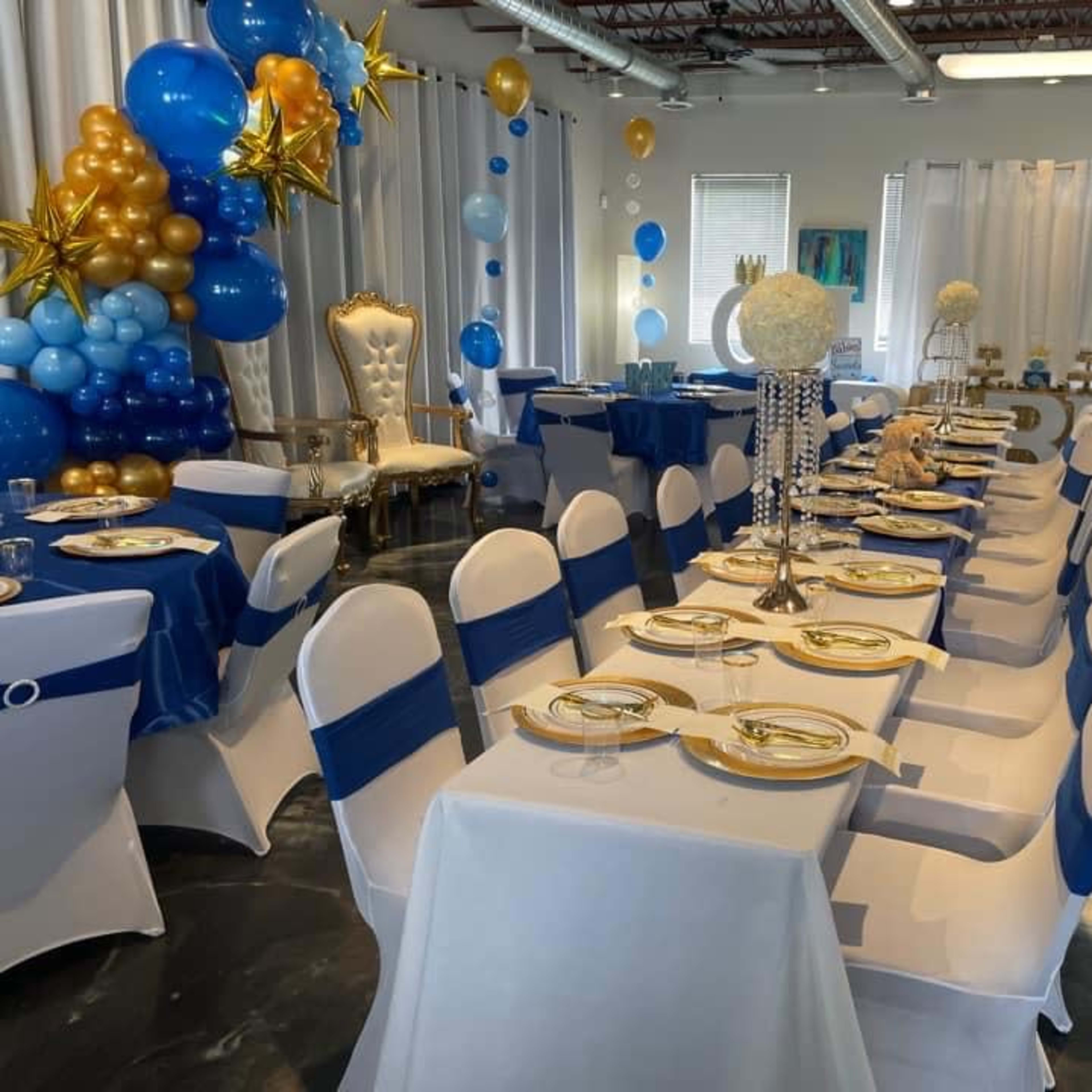 A decorated event space with tables set for a celebration, featuring blue and gold balloons, elegant tablecloths, and a royal throne chair.