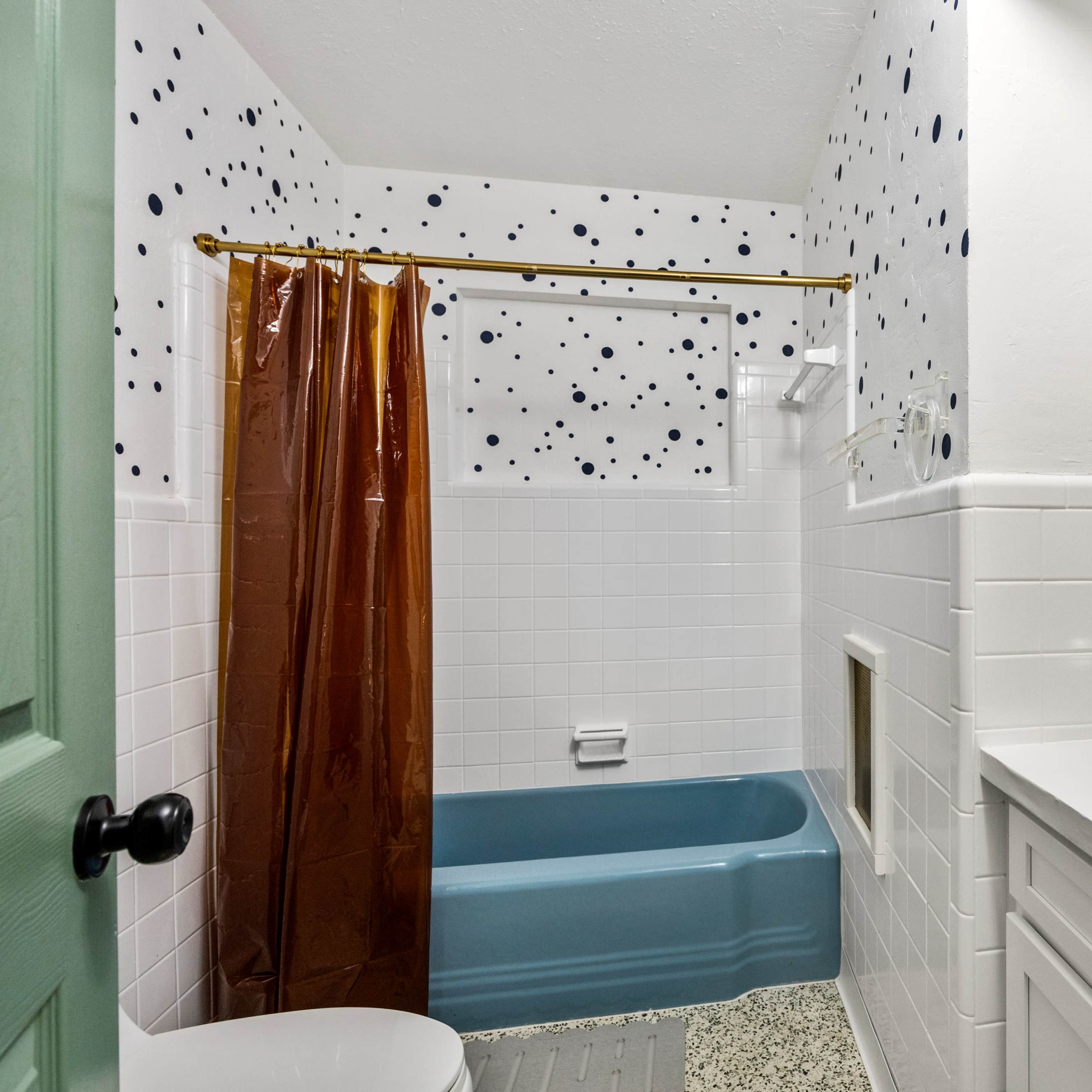 A bathroom features a blue bathtub, orange sink, white tile walls with black dots, and a brown shower curtain.