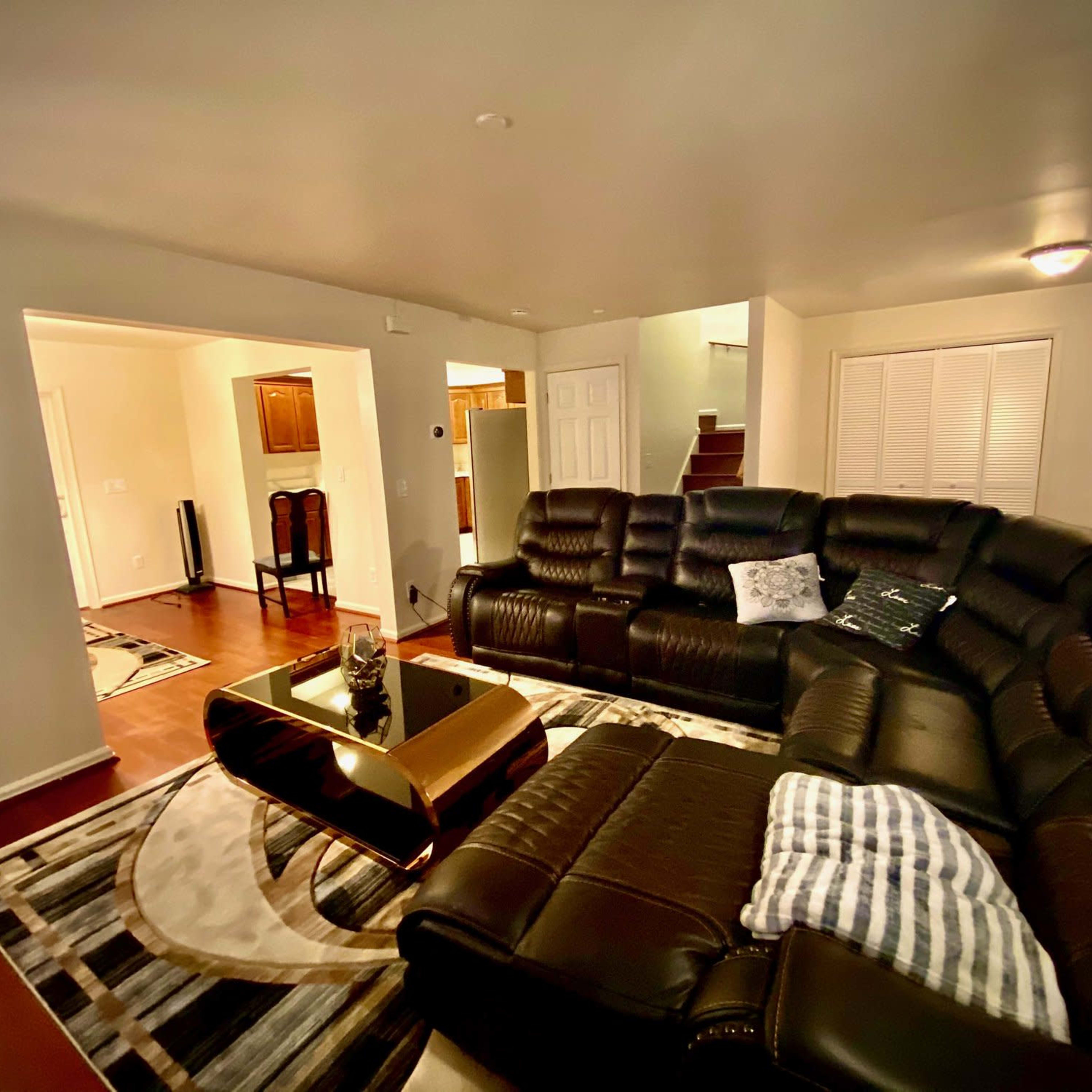A spacious living room featuring a large black sectional sofa, a golden coffee table, and a patterned rug on the wooden floor.