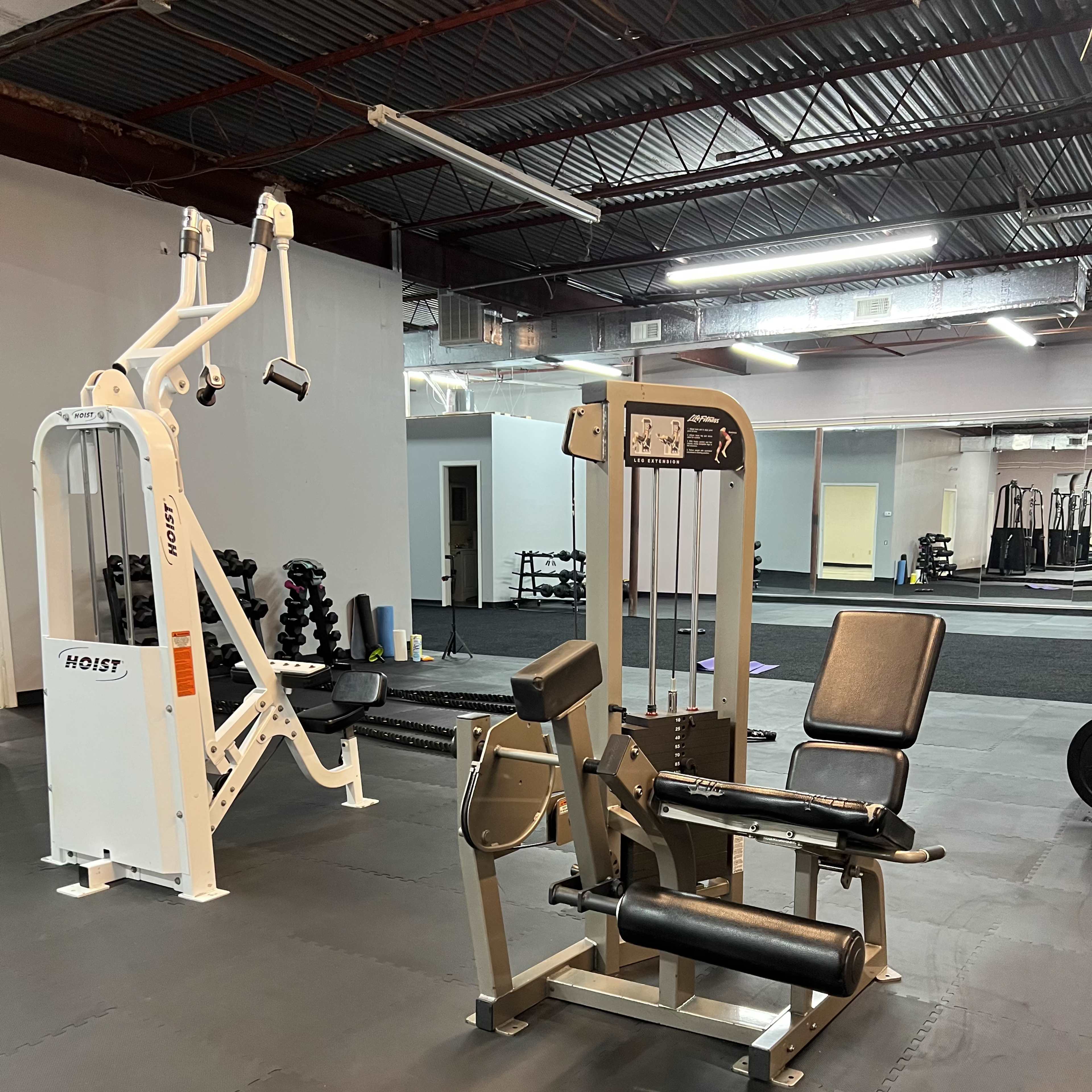 A fitness center interior featuring weight machines, including a Hoist machine on the left and a leg extension machine on the right, set on black rubber flooring.