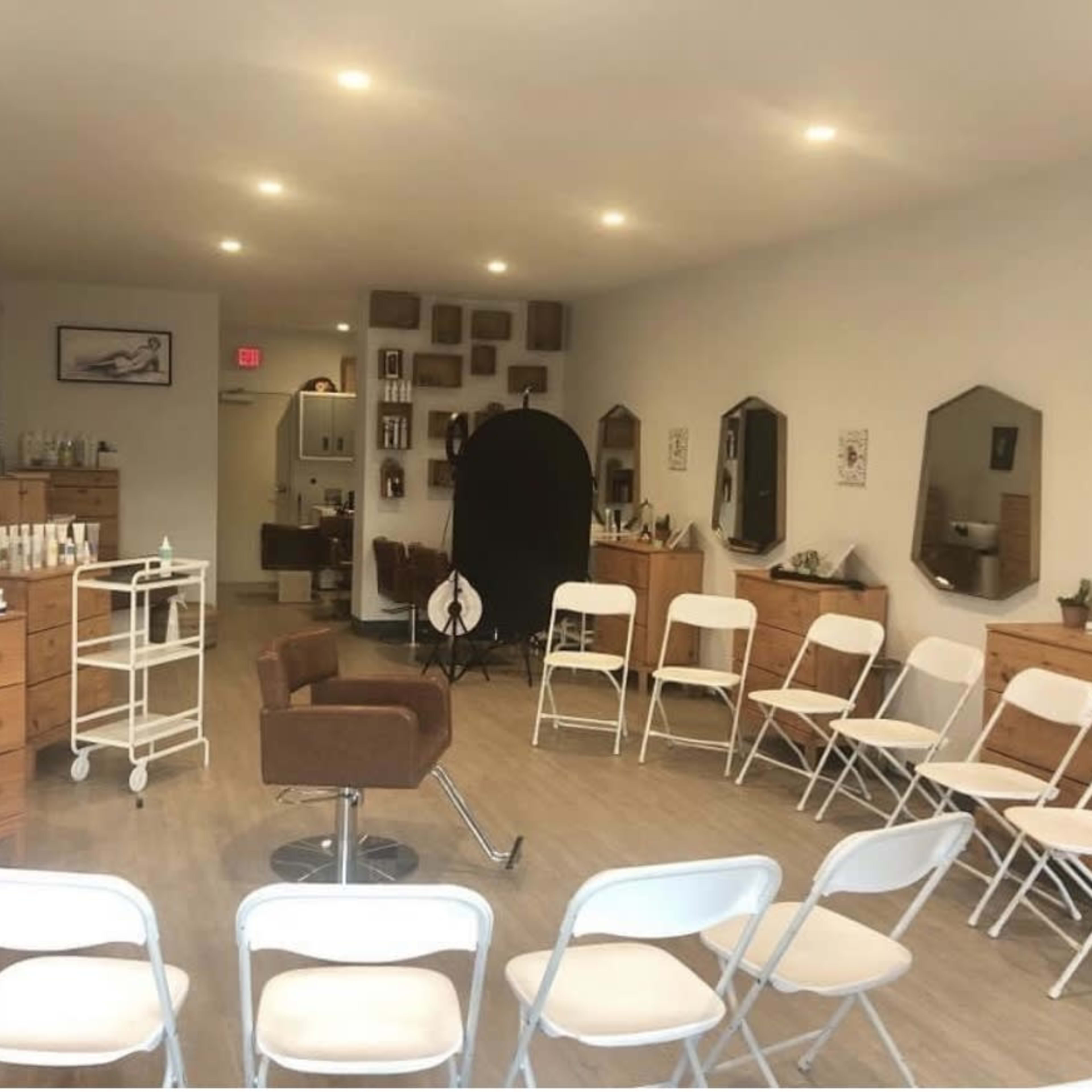 A hair salon interior with wooden dressers, a styling chair in the center, and white folding chairs arranged in a semi-circle.