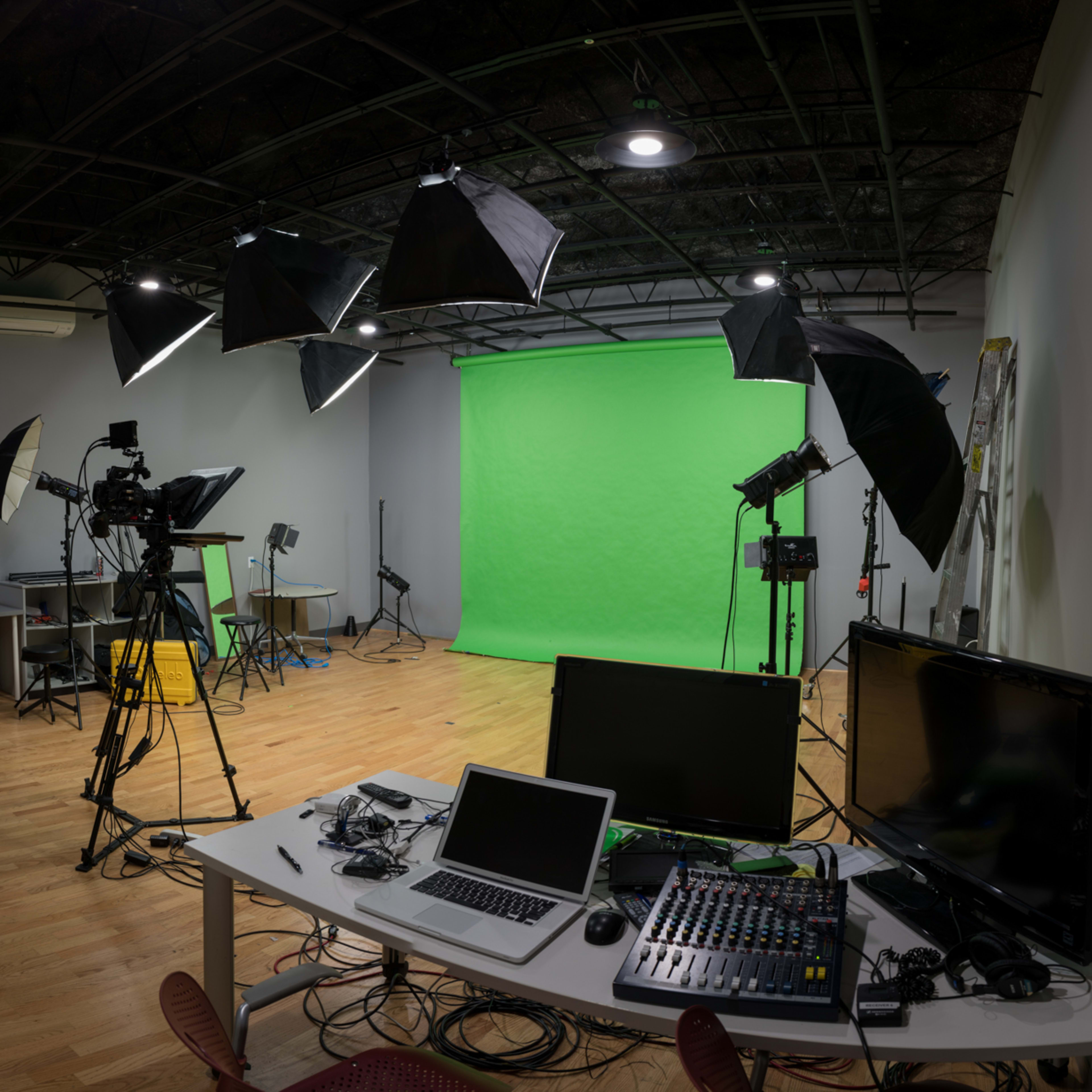 A studio setup featuring a green screen, multiple lights on stands, cameras, and a desk with computer equipment and sound mixing gear.