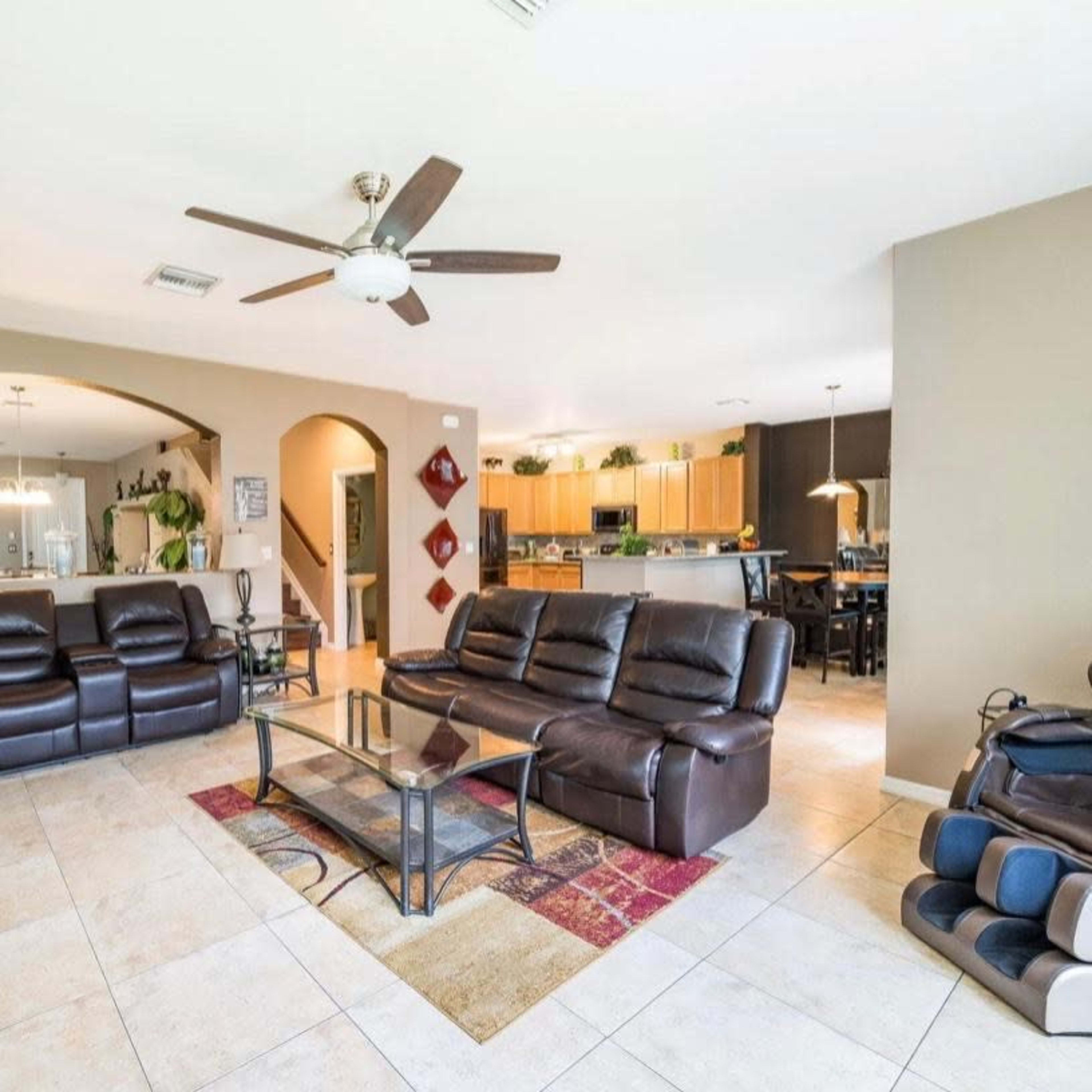 A spacious living room features dark leather sofas, a glass coffee table, and a TV, with an open view to a kitchen area in the background.