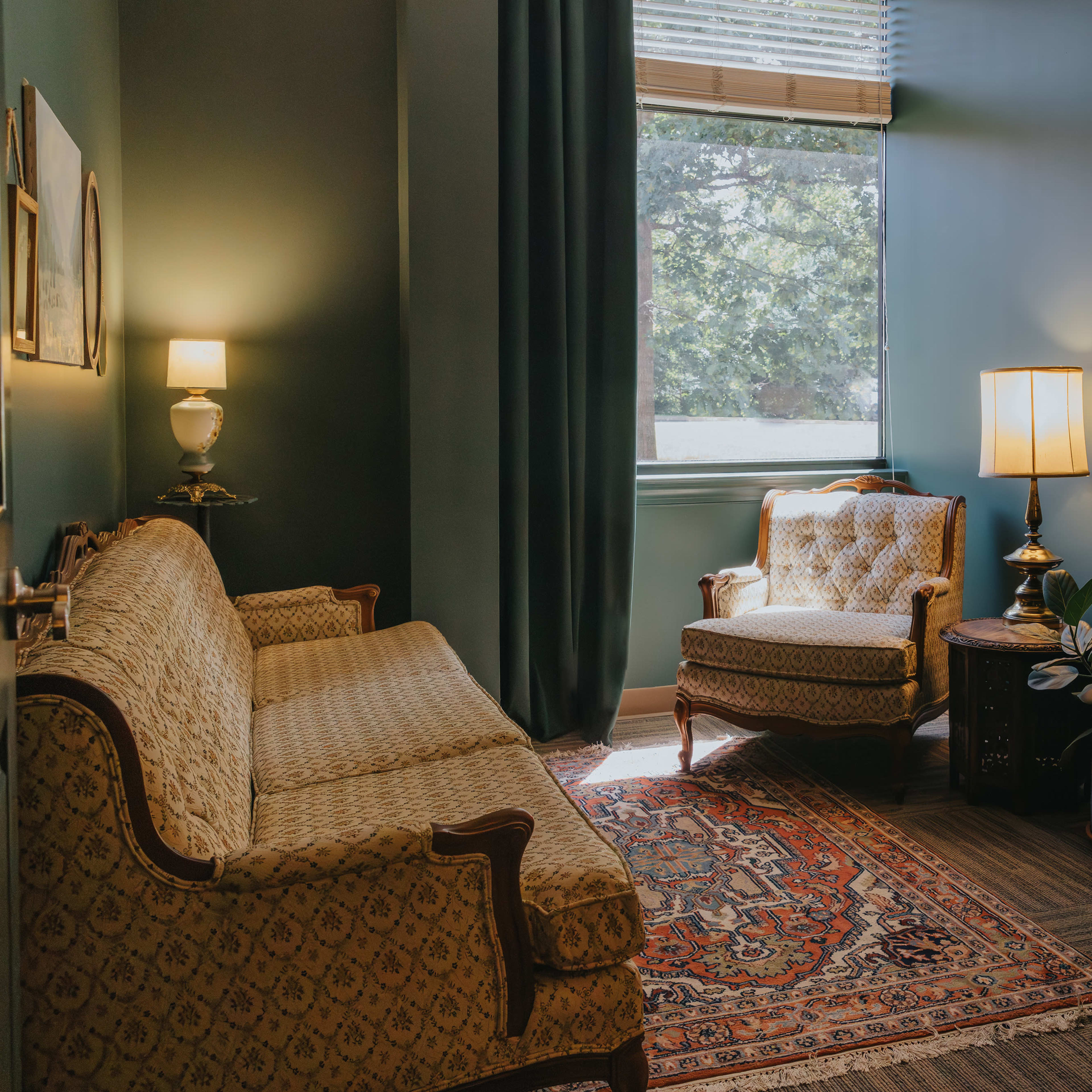 A cozy room featuring a patterned sofa, a vintage armchair, a wooden desk, and a large window allowing natural light to illuminate the space.