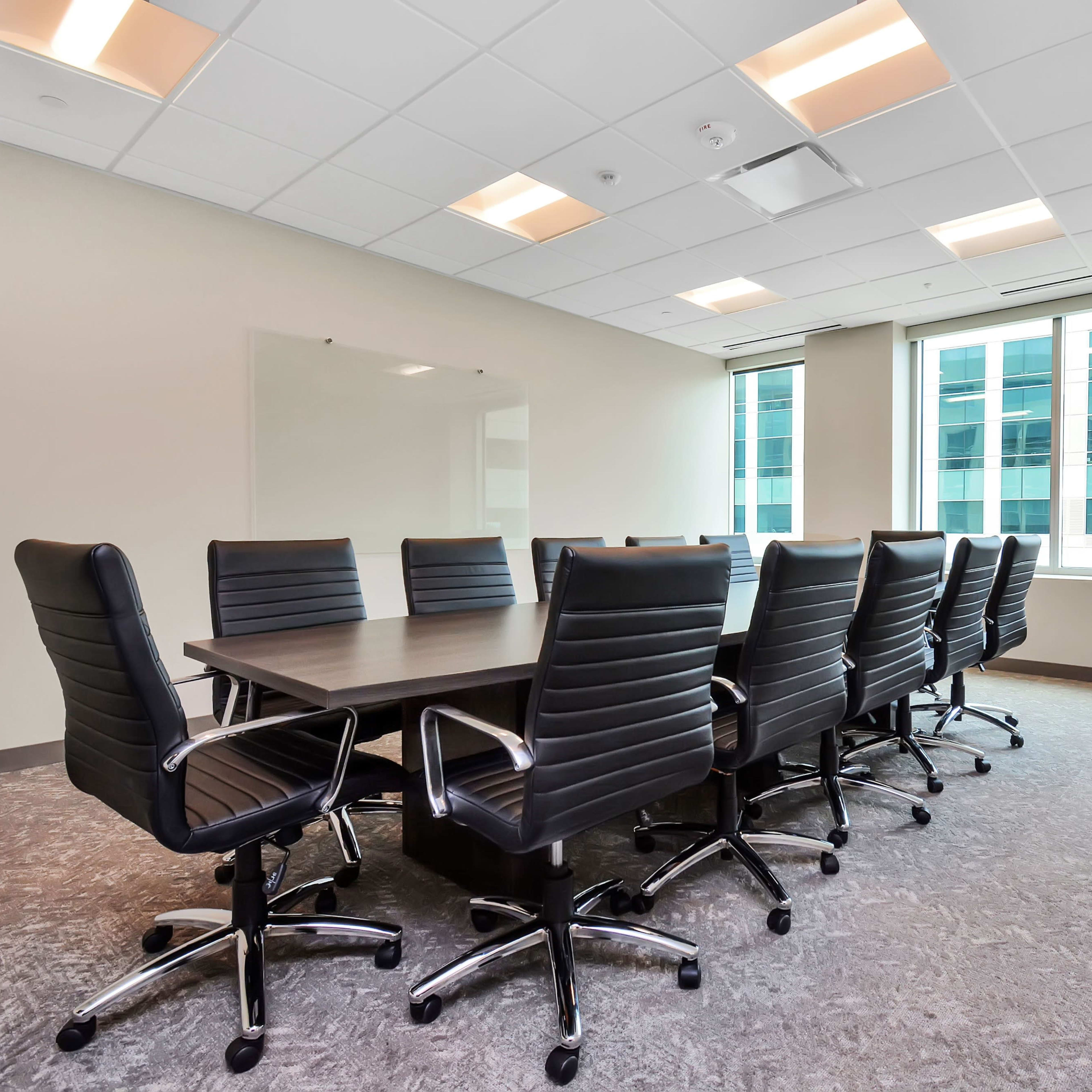 A large conference room features a rectangular table surrounded by eight black swivel chairs, with windows overlooking an urban landscape.