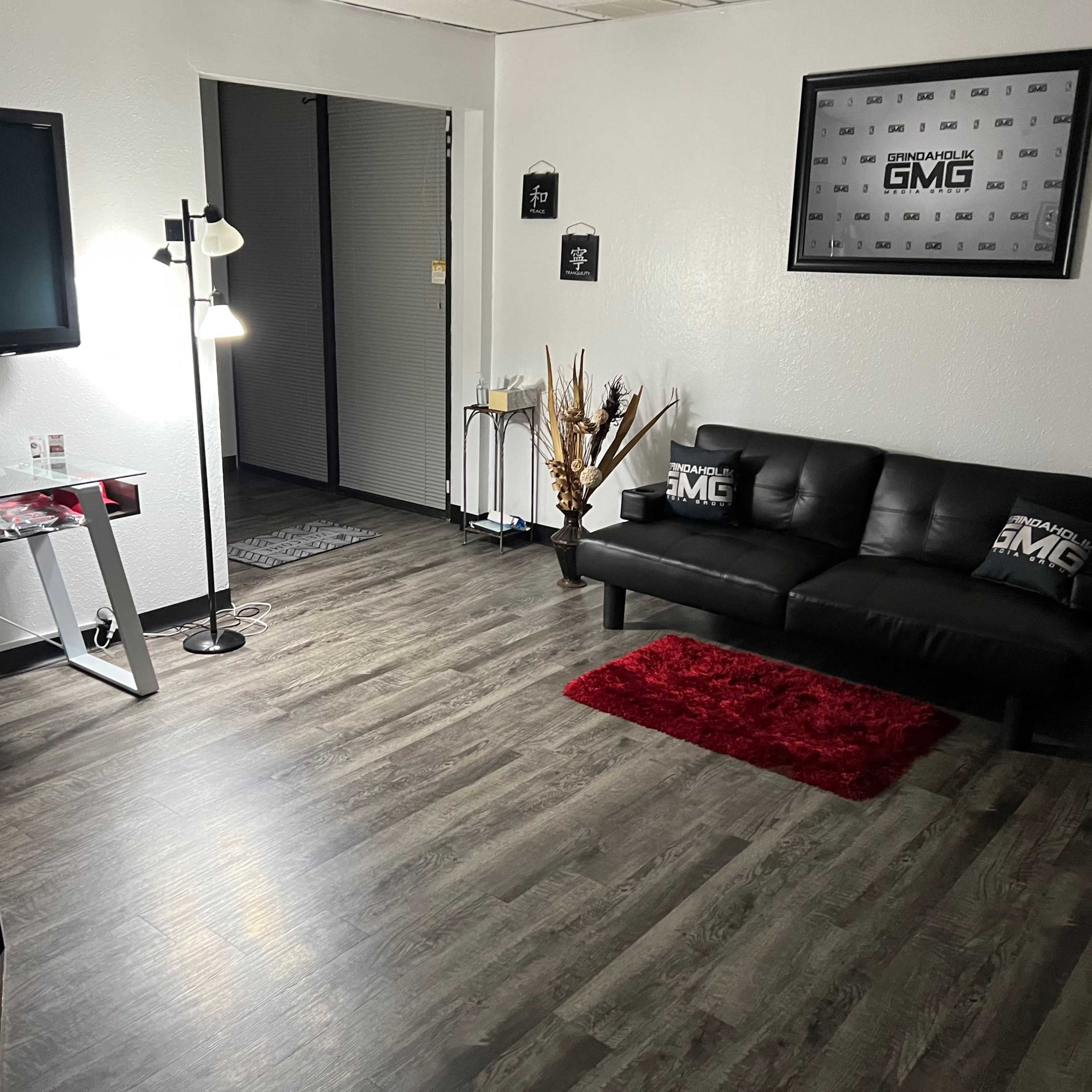 A modern office space with a black couch, a television, and a lamp, featuring a wooden floor and a red rug.