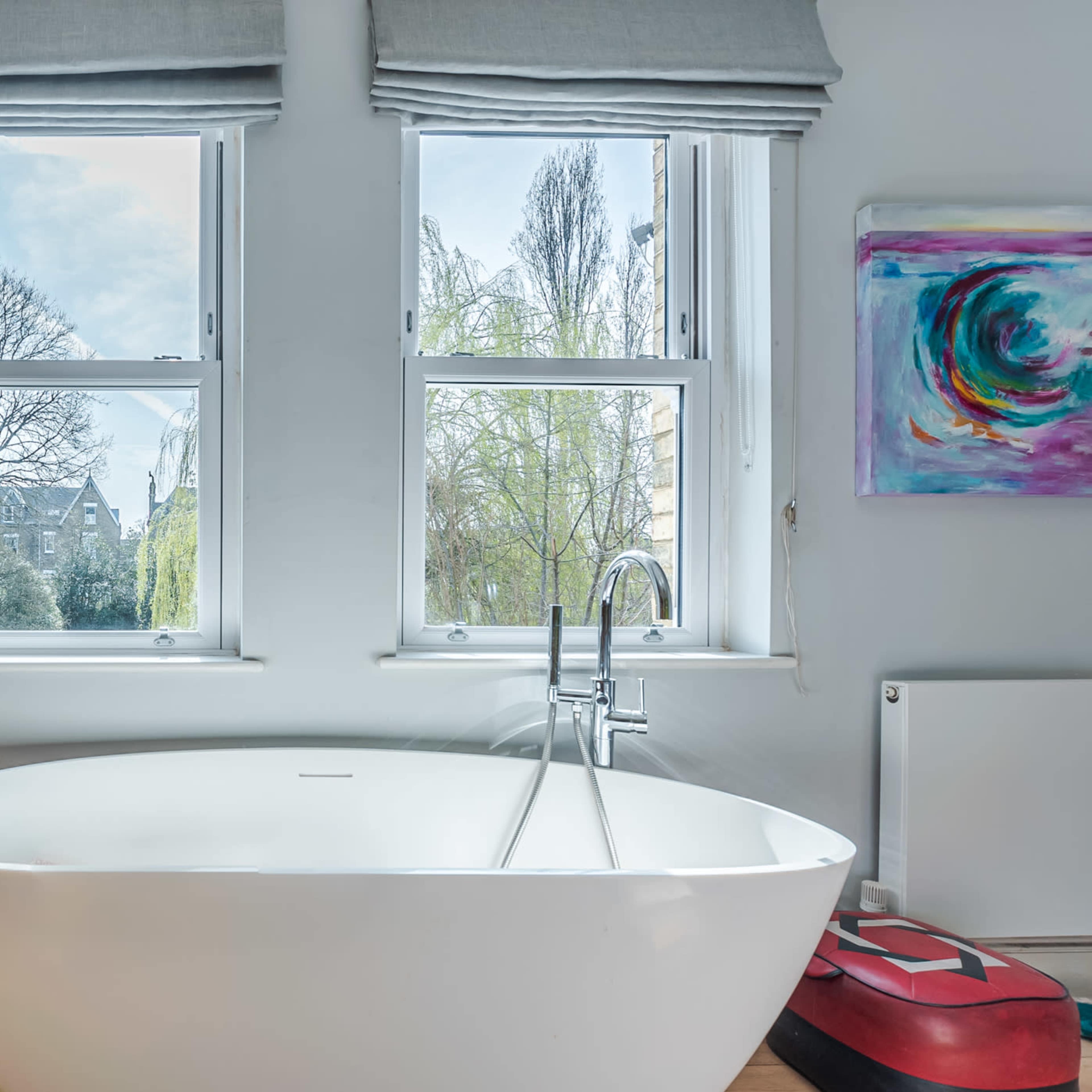 A modern bathroom featuring a freestanding white bathtub positioned between two windows, with a wooden storage cabinet nearby and a colorful abstract painting on the wall.
