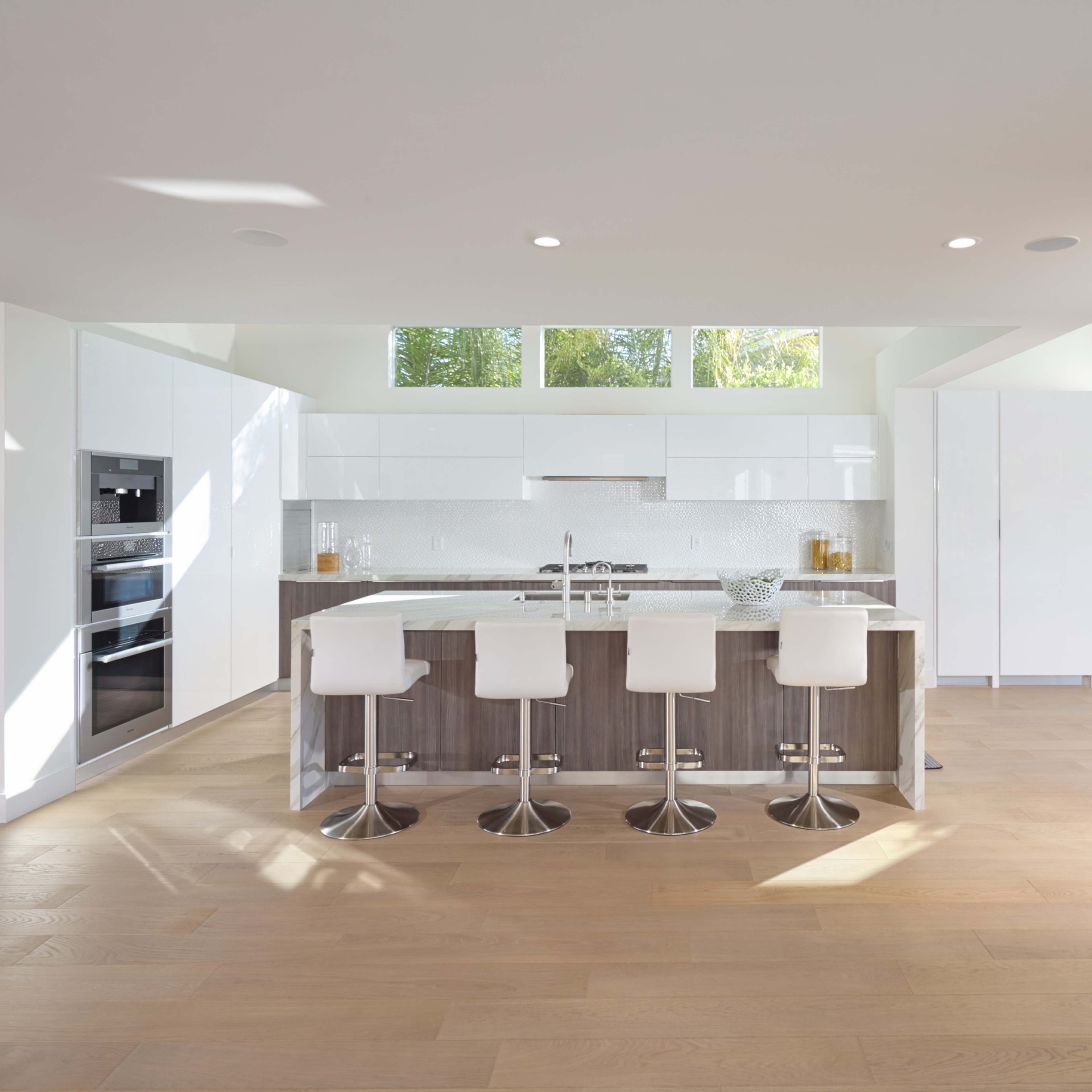 A modern kitchen with a central island, four white bar stools, and large windows allowing natural light to fill the space.