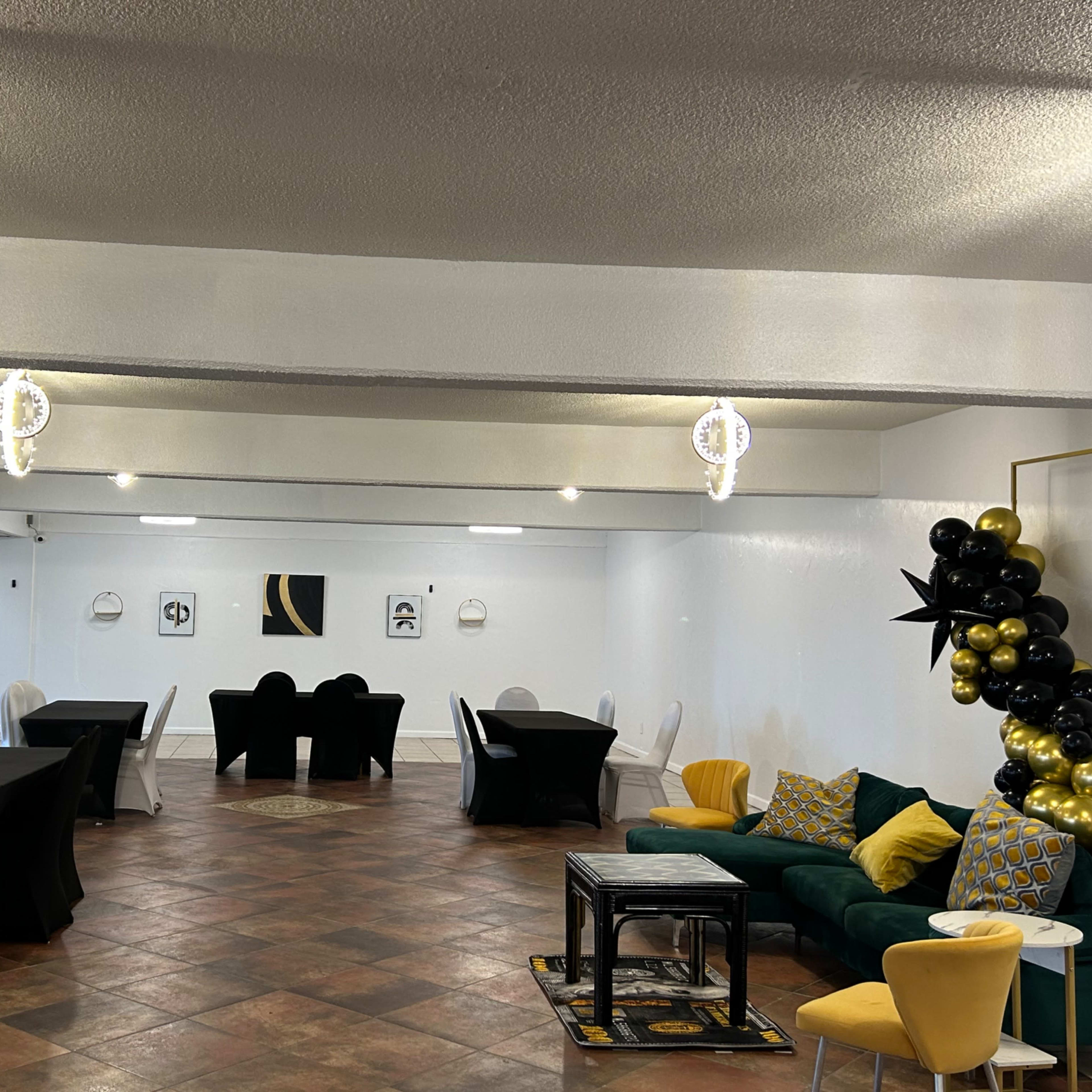 A spacious event room featuring black and white tables, a green sofa with decorative cushions, and balloon decorations along with modern lighting fixtures.