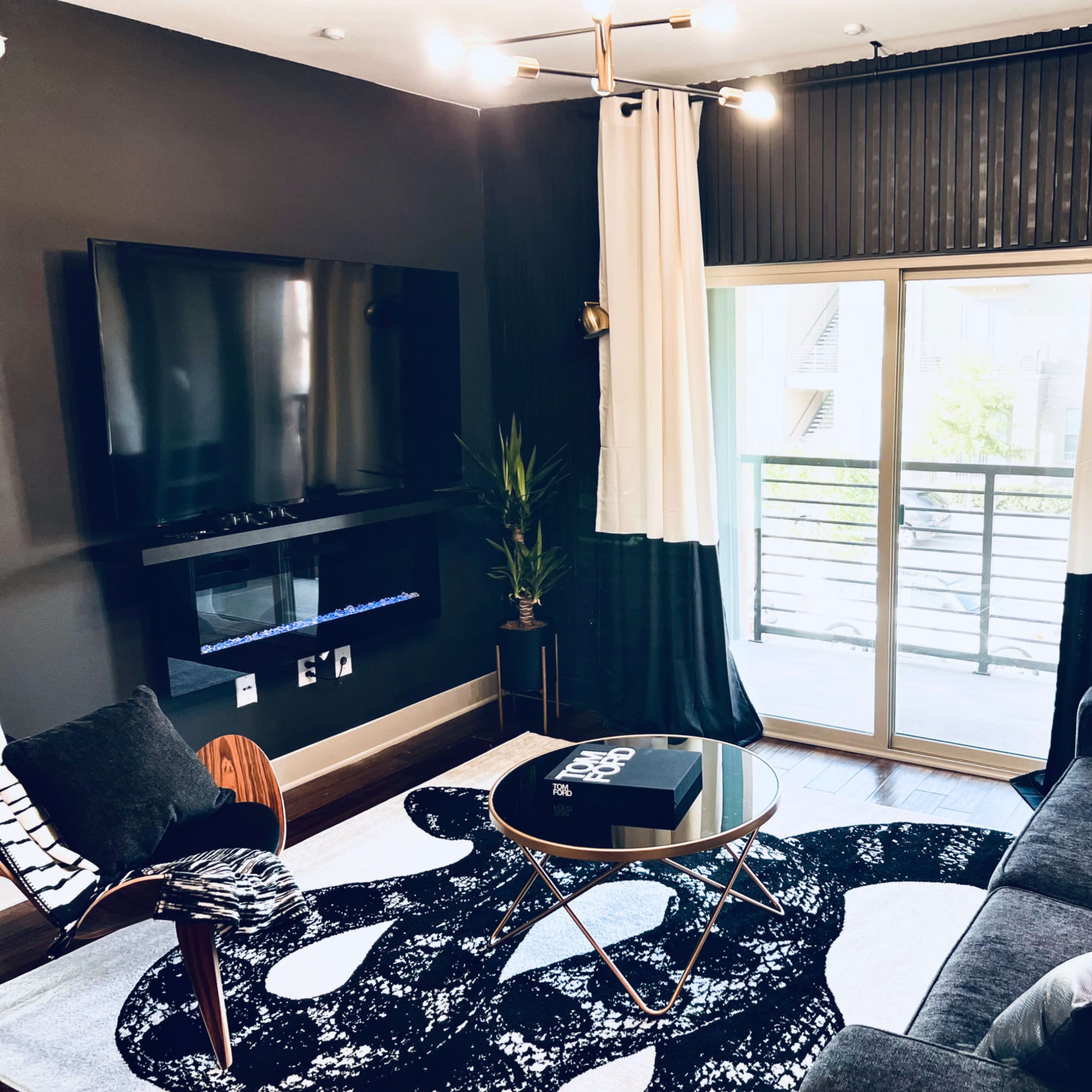 The living room features a dark wall, a large TV mounted above a low console, a comfortable sofa, and a round coffee table on a patterned rug, with large windows allowing natural light.