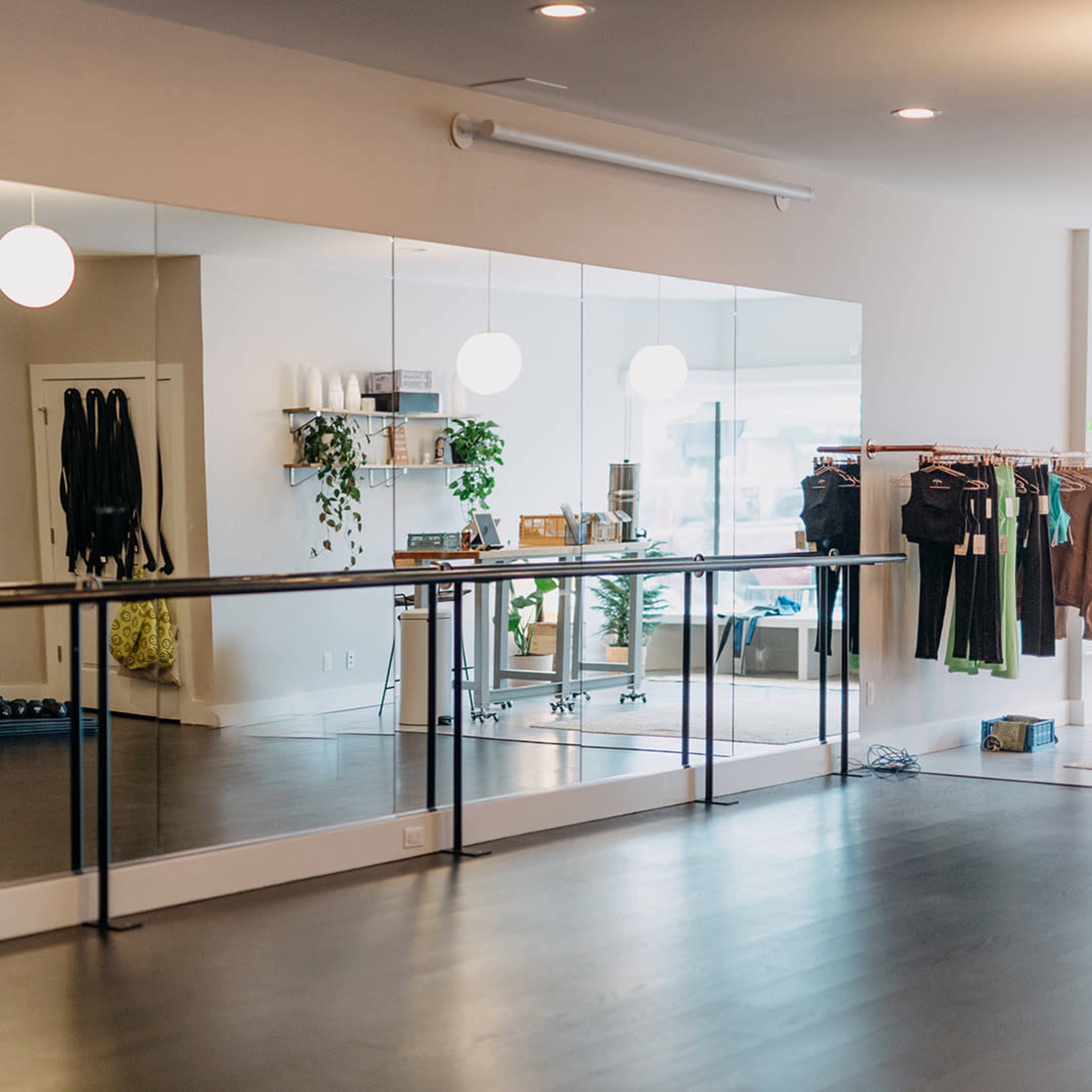 A fitness studio is equipped with large mirrors, exercise mats, dumbbells, and clothing racks along the wall.