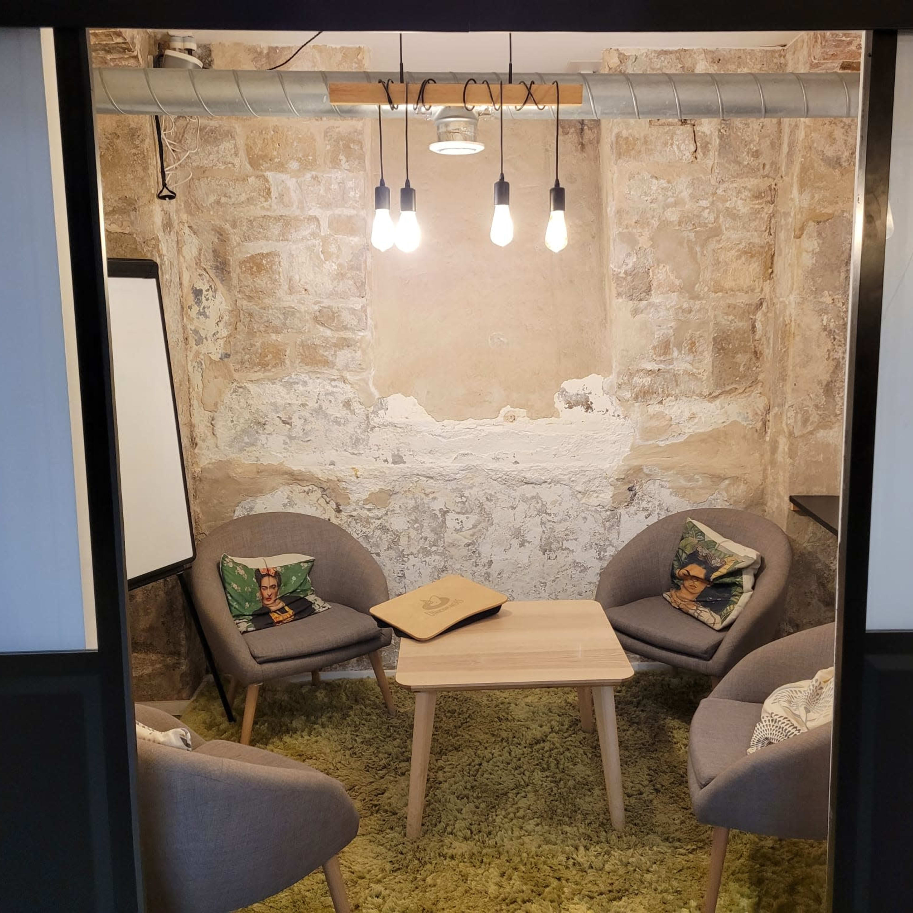 A small meeting room with four gray chairs arranged around a wooden table, illuminated by hanging light bulbs against a stone wall.