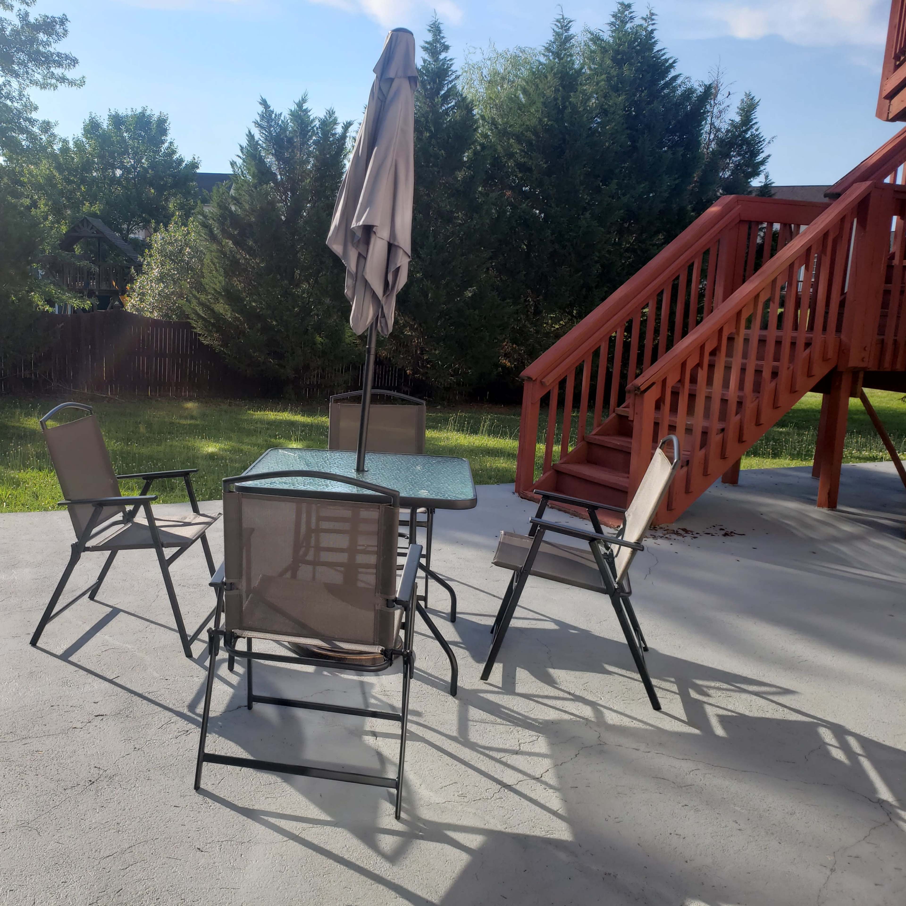 A patio area features a glass table surrounded by four metal chairs, with a large umbrella and a wooden deck in the background.
