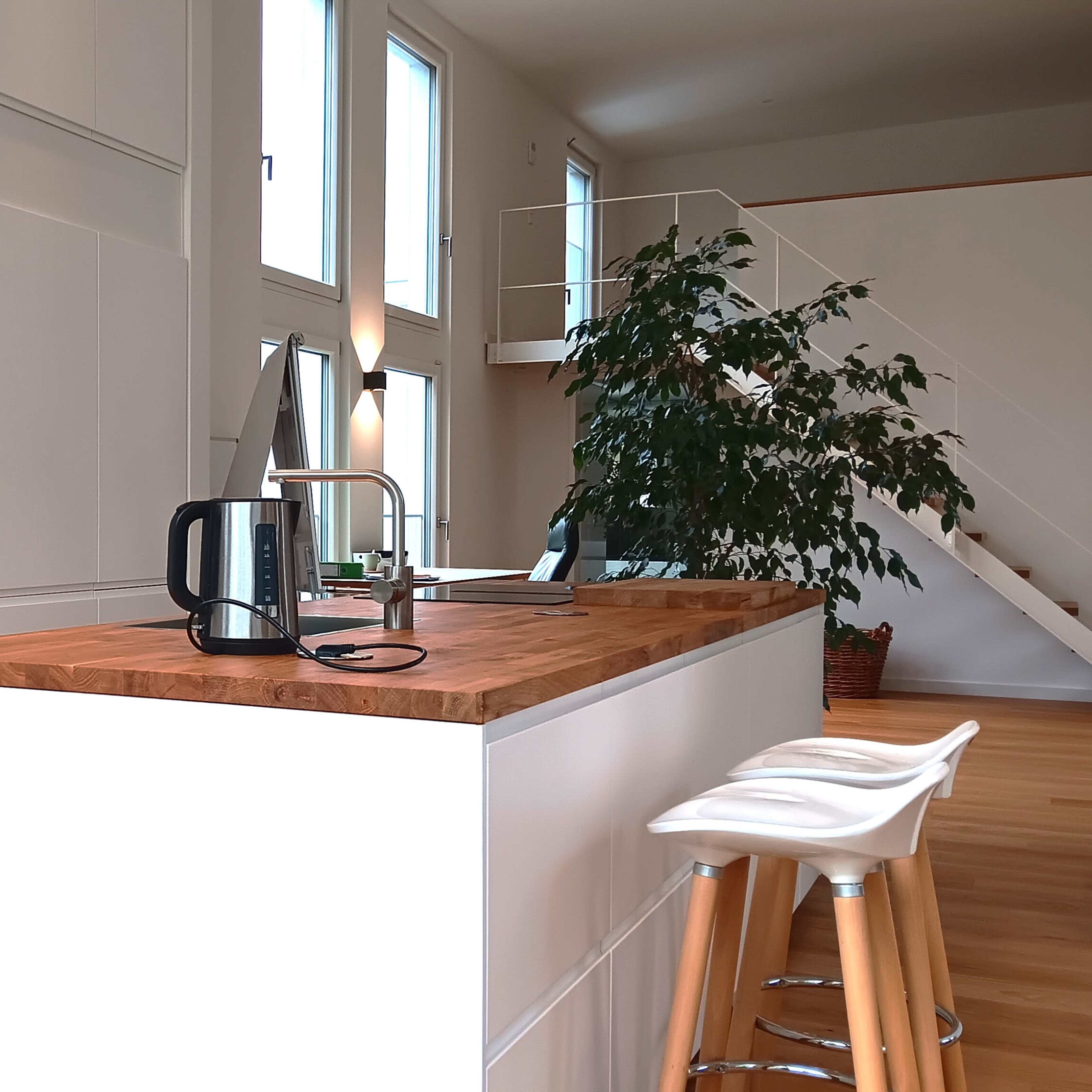 A bright kitchen with white cabinetry, a wooden countertop island, a kettle, and a staircase in the background.