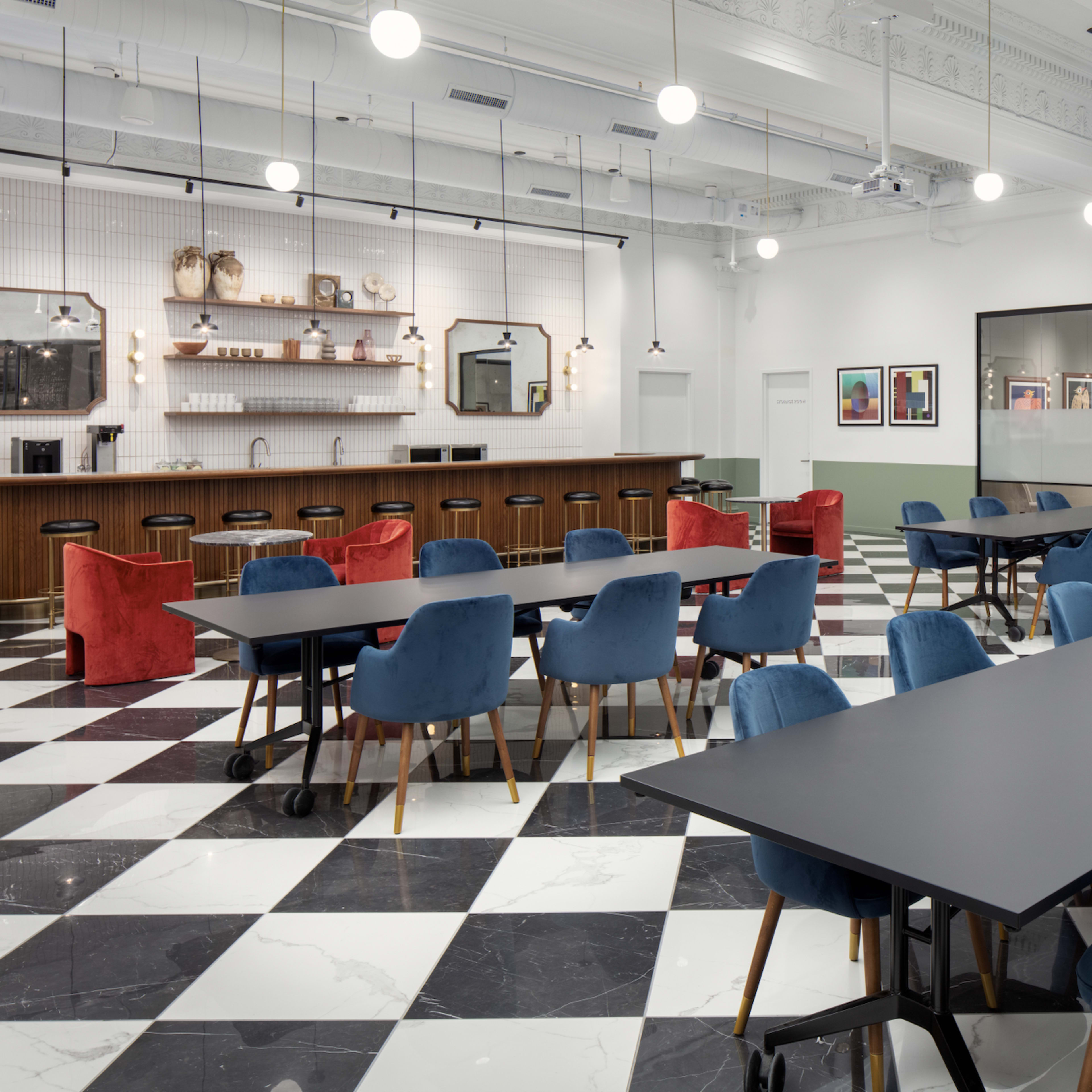 A modern, spacious dining area with checkered black and white flooring, featuring several tables surrounded by blue and red chairs, and a bar counter in the background.