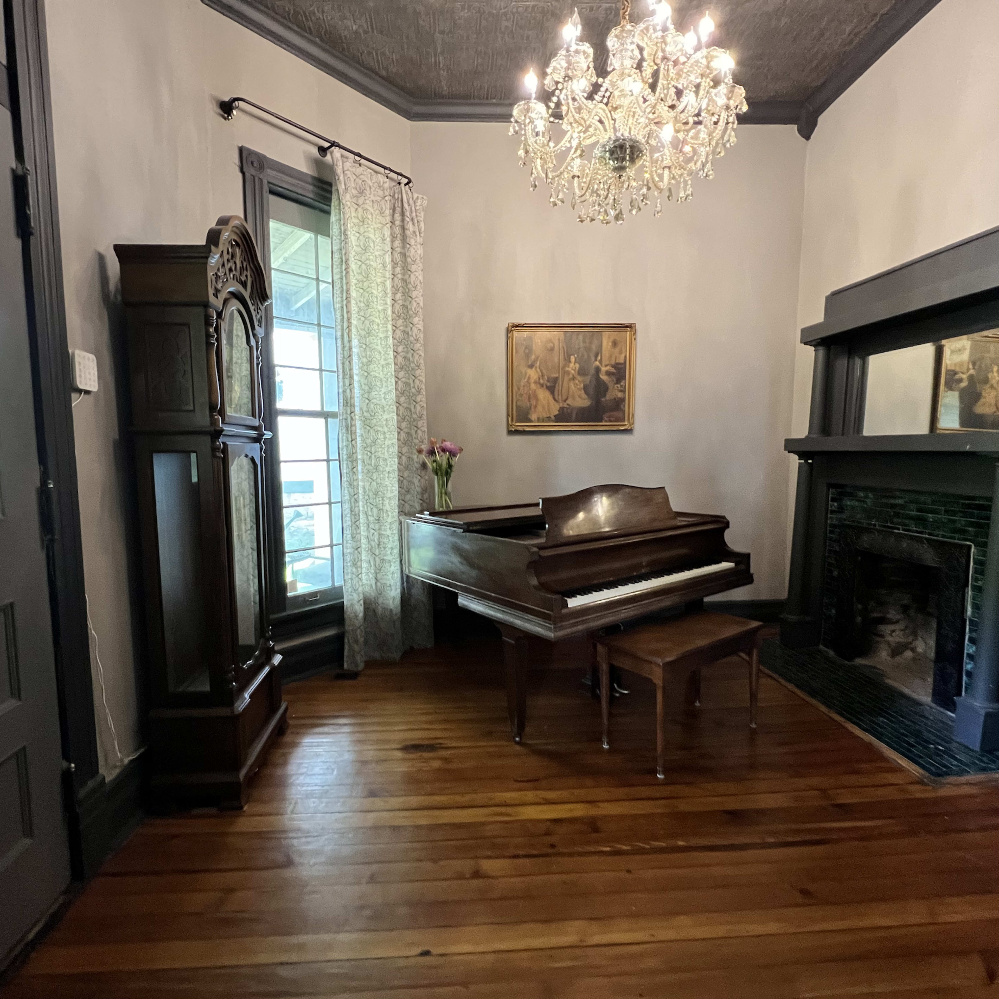 A grand piano and a small stool are positioned beside a fireplace in a room with wooden flooring and a chandelier overhead.