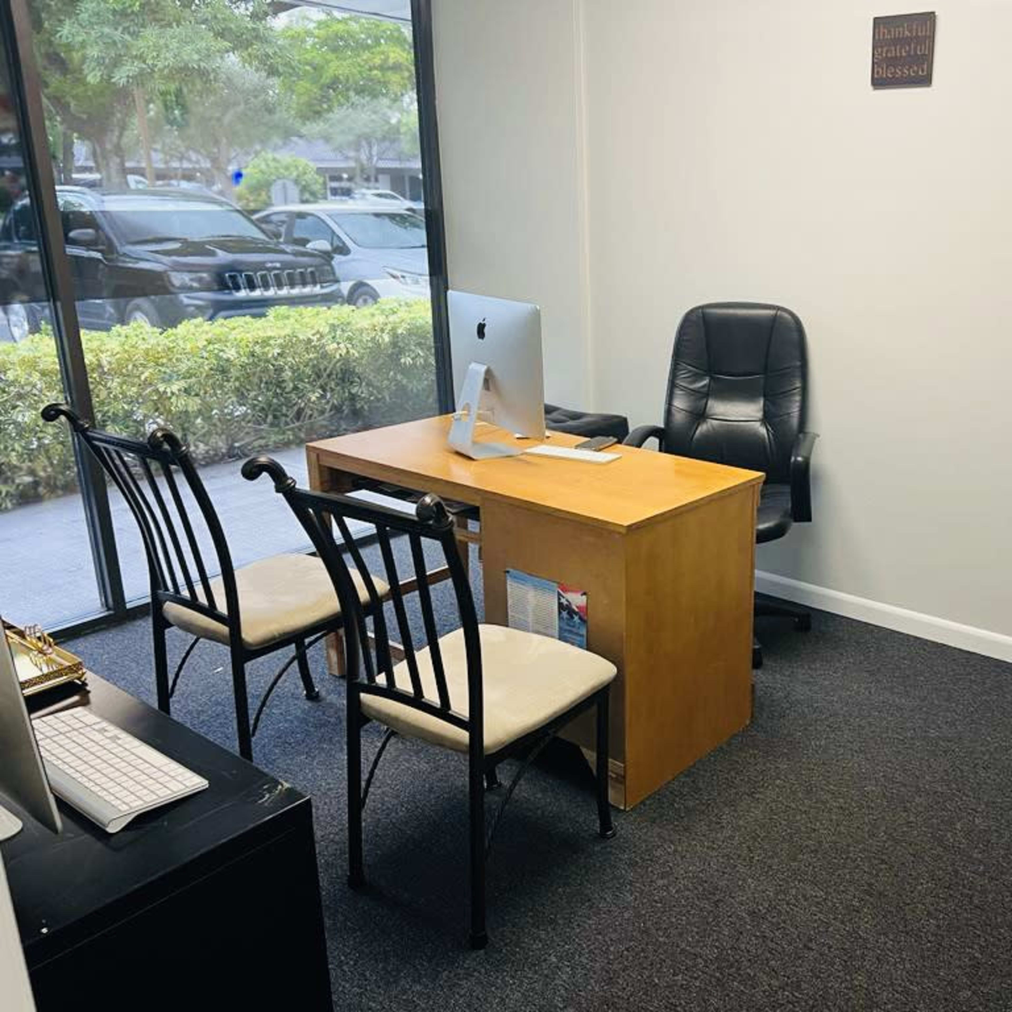 A small office space with a wooden desk, a black ergonomic chair, two empty chairs, and a large window facing a landscaped area.