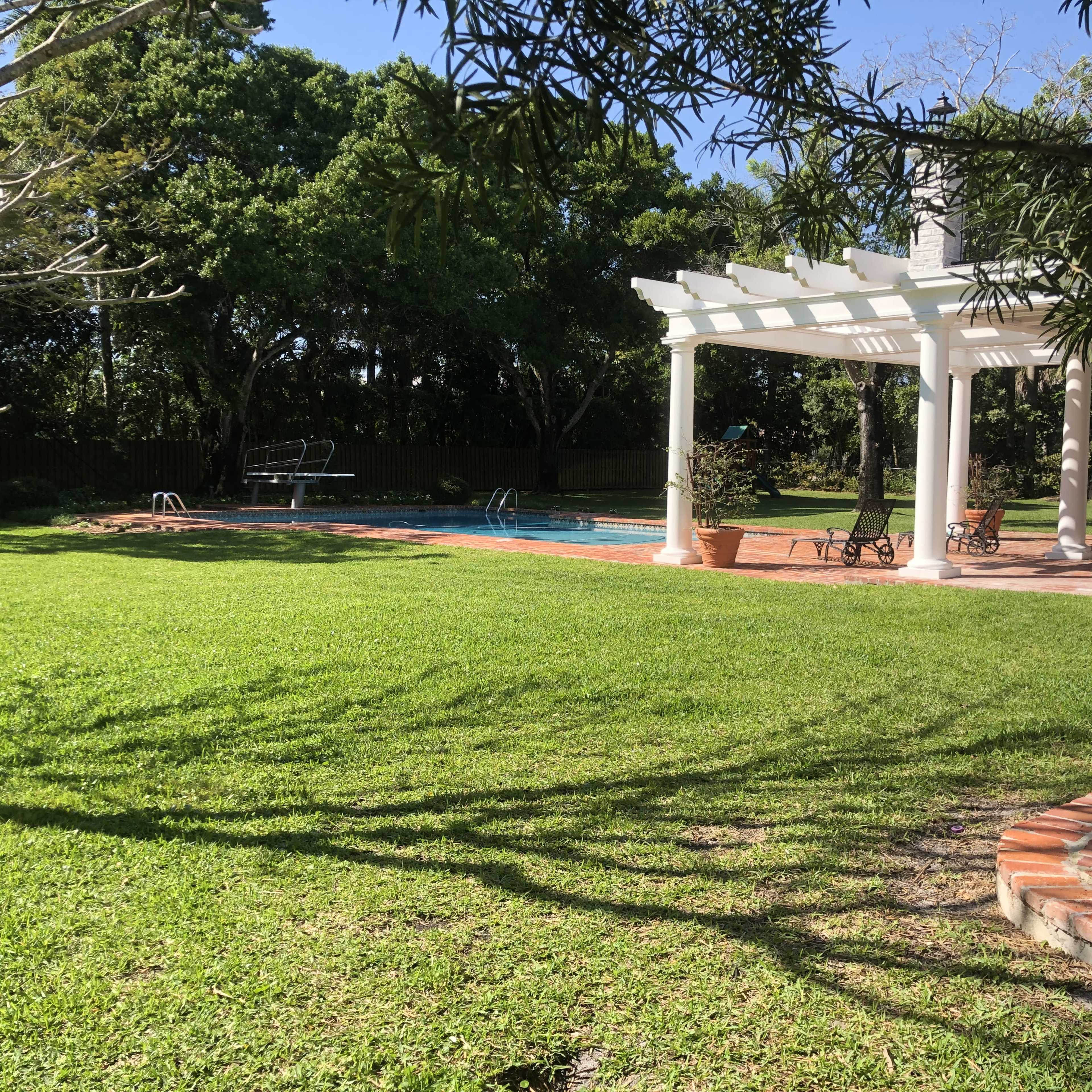 A pool area surrounded by a well-maintained lawn and a pergola with chairs nearby.