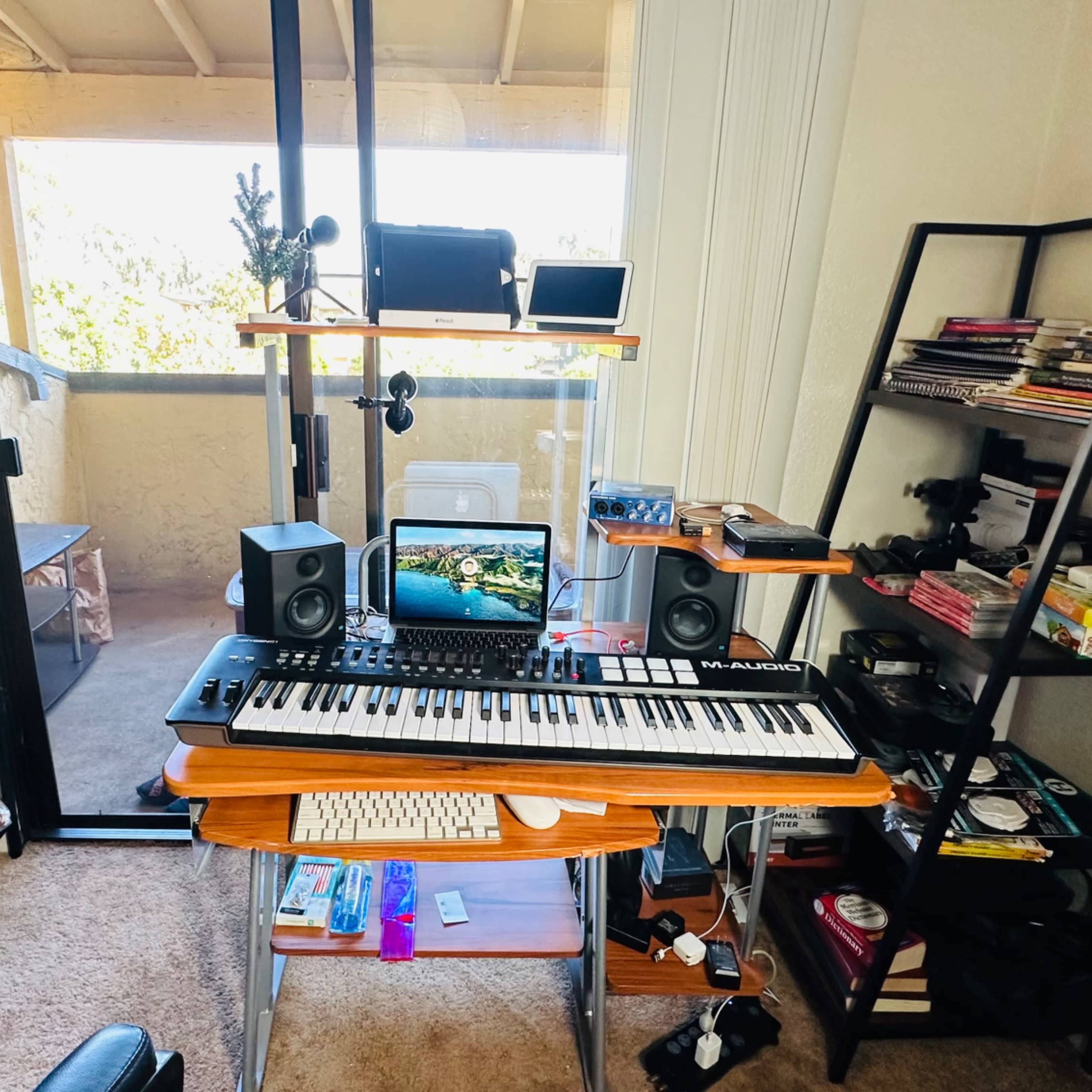 A home office setup features a keyboard on a desk with speakers and a computer monitor, surrounded by shelves filled with books and various equipment.
