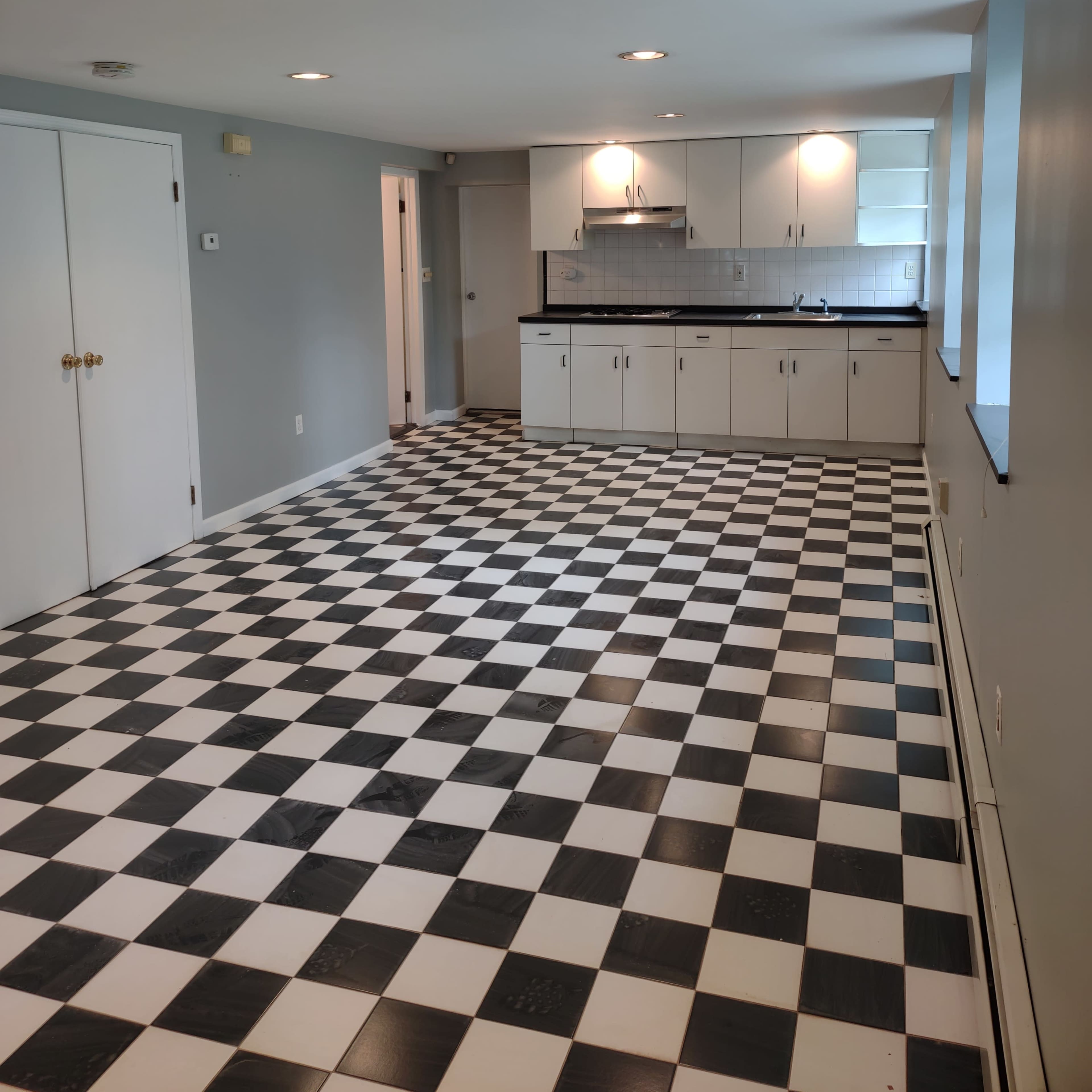 A spacious, open room featuring a black and white checkered floor and a kitchen area with white cabinets and a black countertop.