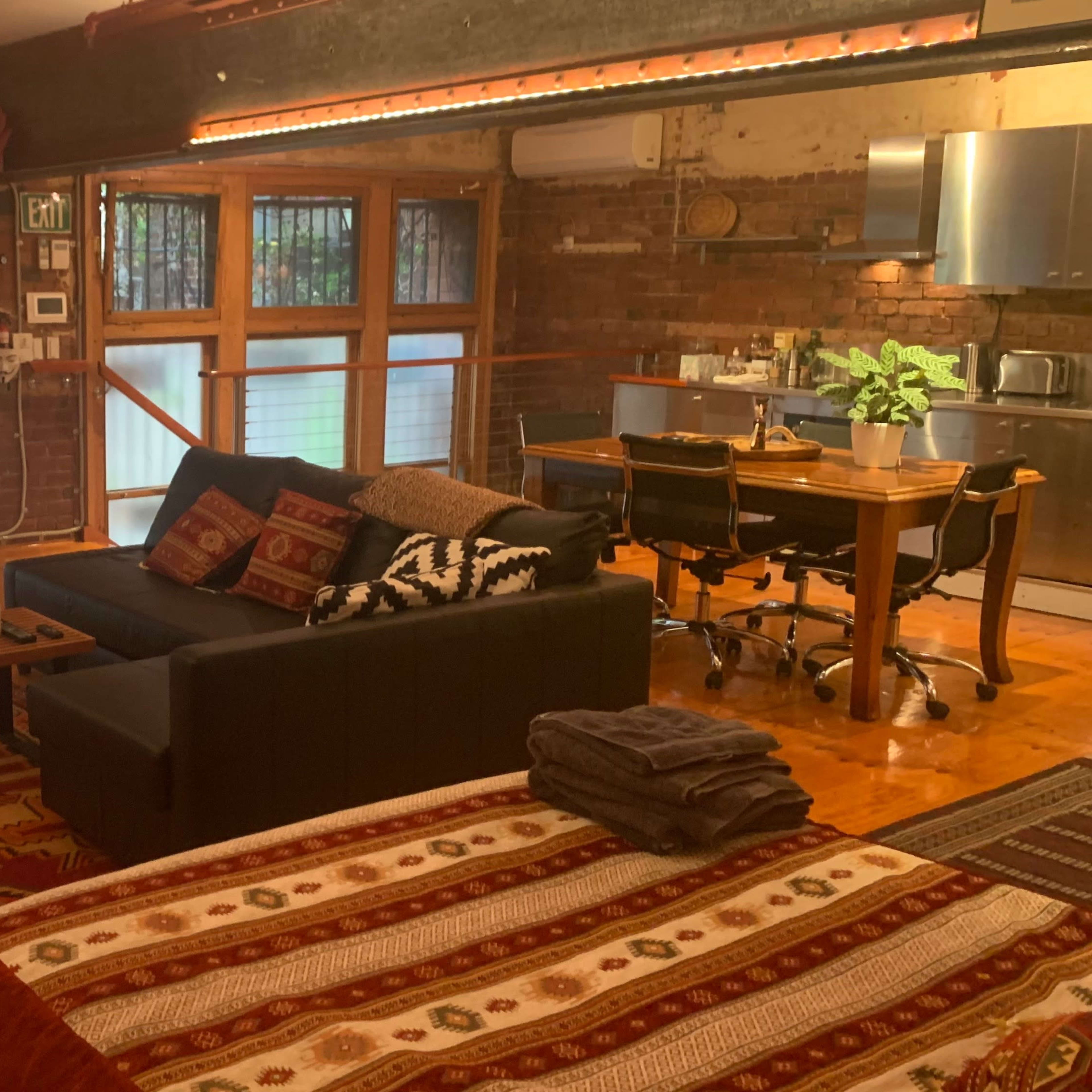 A stylish loft with exposed brick walls, hardwood floors, and a living area featuring a sofa, a dining table, and a workspace beside large windows.