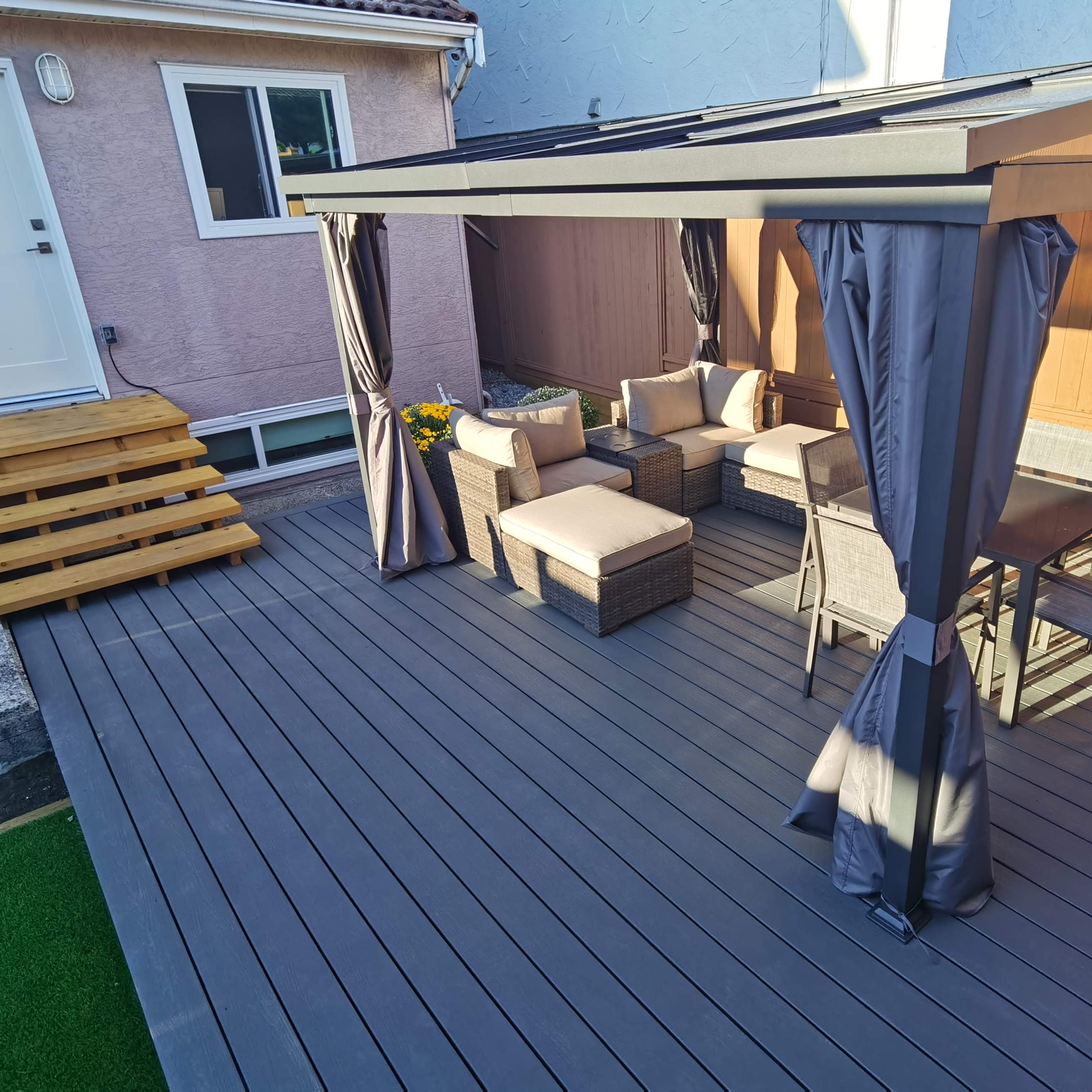 A patio area features a covered seating arrangement with gray curtains, a table, and chairs, surrounded by wooden decking and a small set of steps leading to a door.