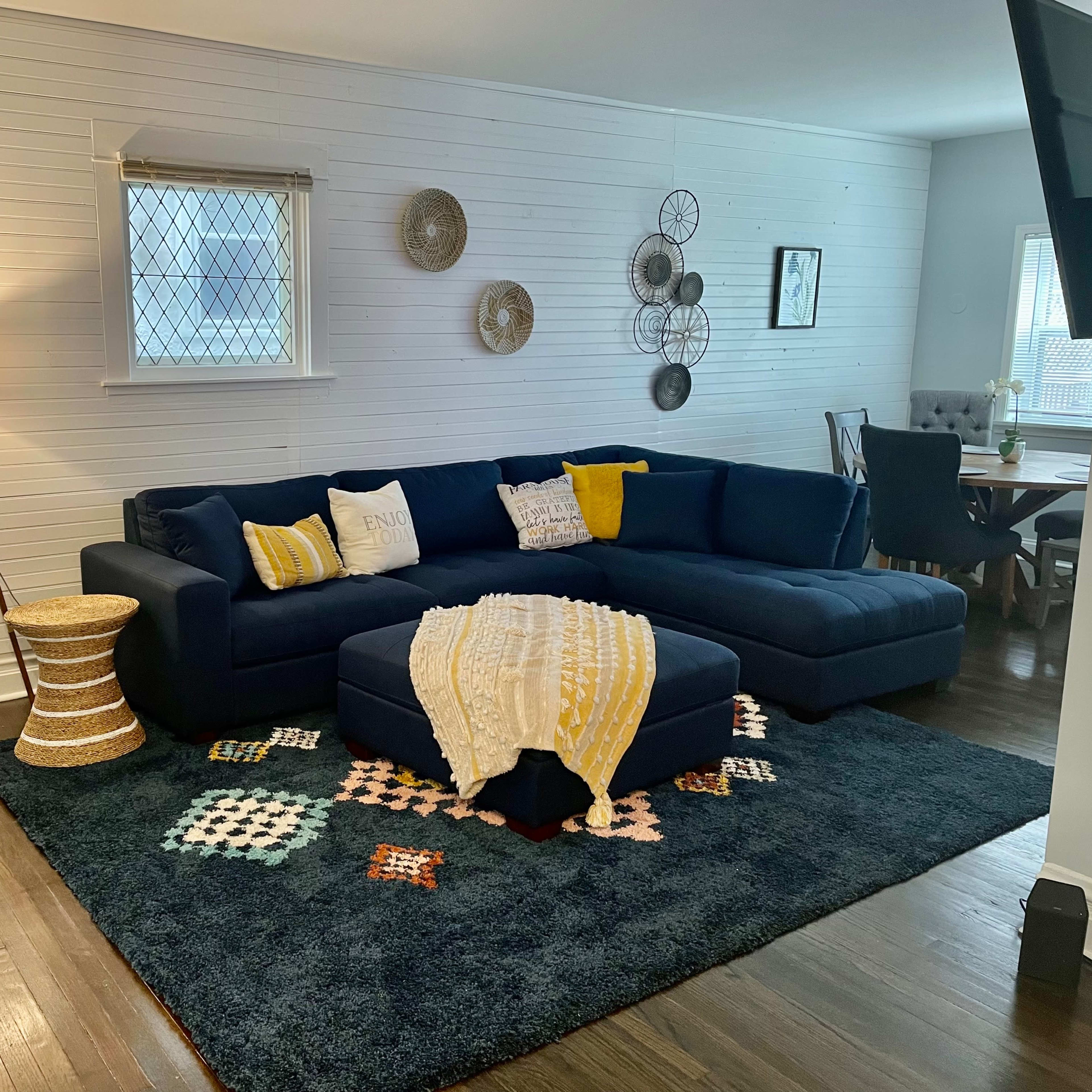 A living room with a dark blue sectional sofa, a round mirror on the wall, a lamp on a tripod stand, and a dining area in the background.