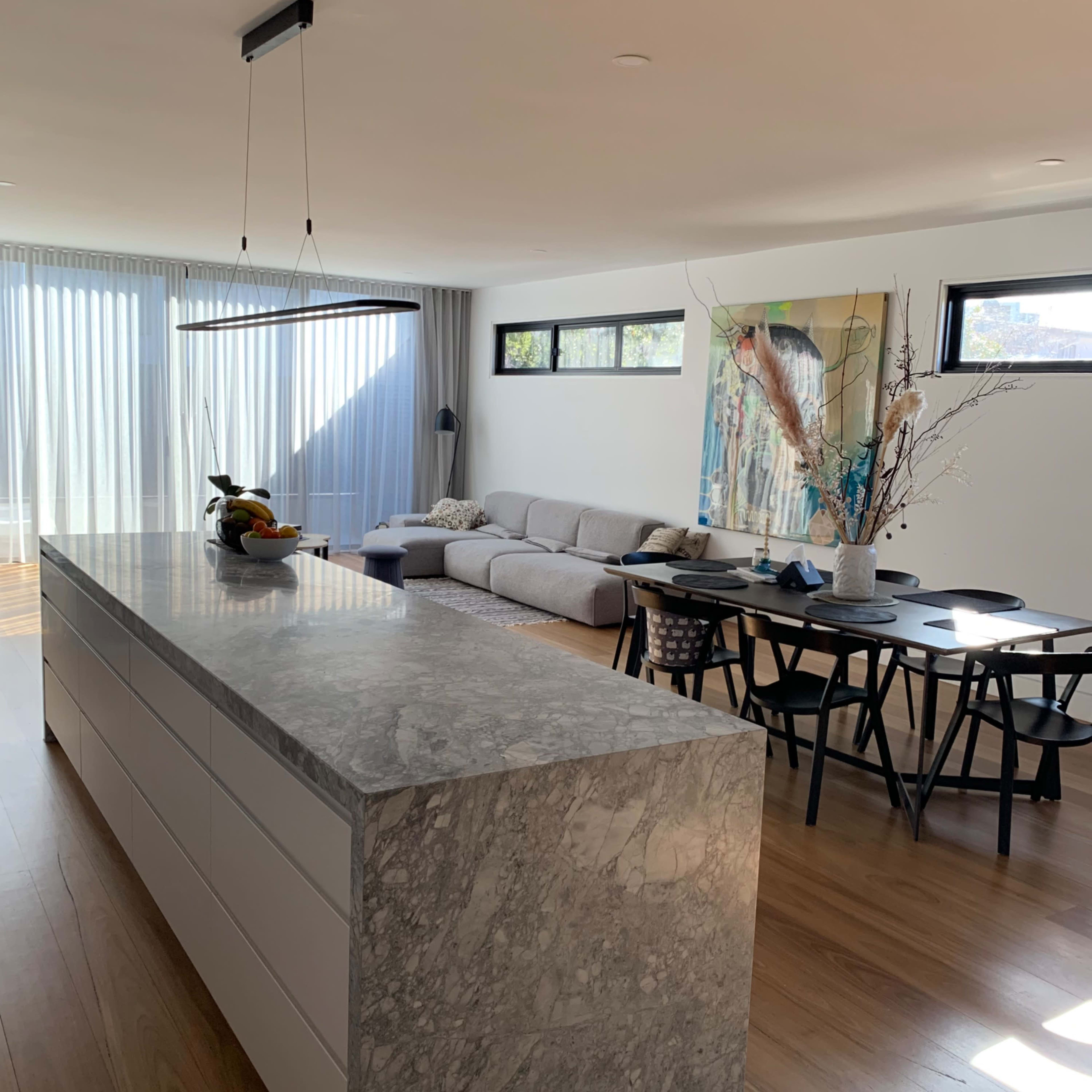 A modern open-concept kitchen and living area features a stone island, a grey sofa, a dining table, and large windows with sheer curtains.