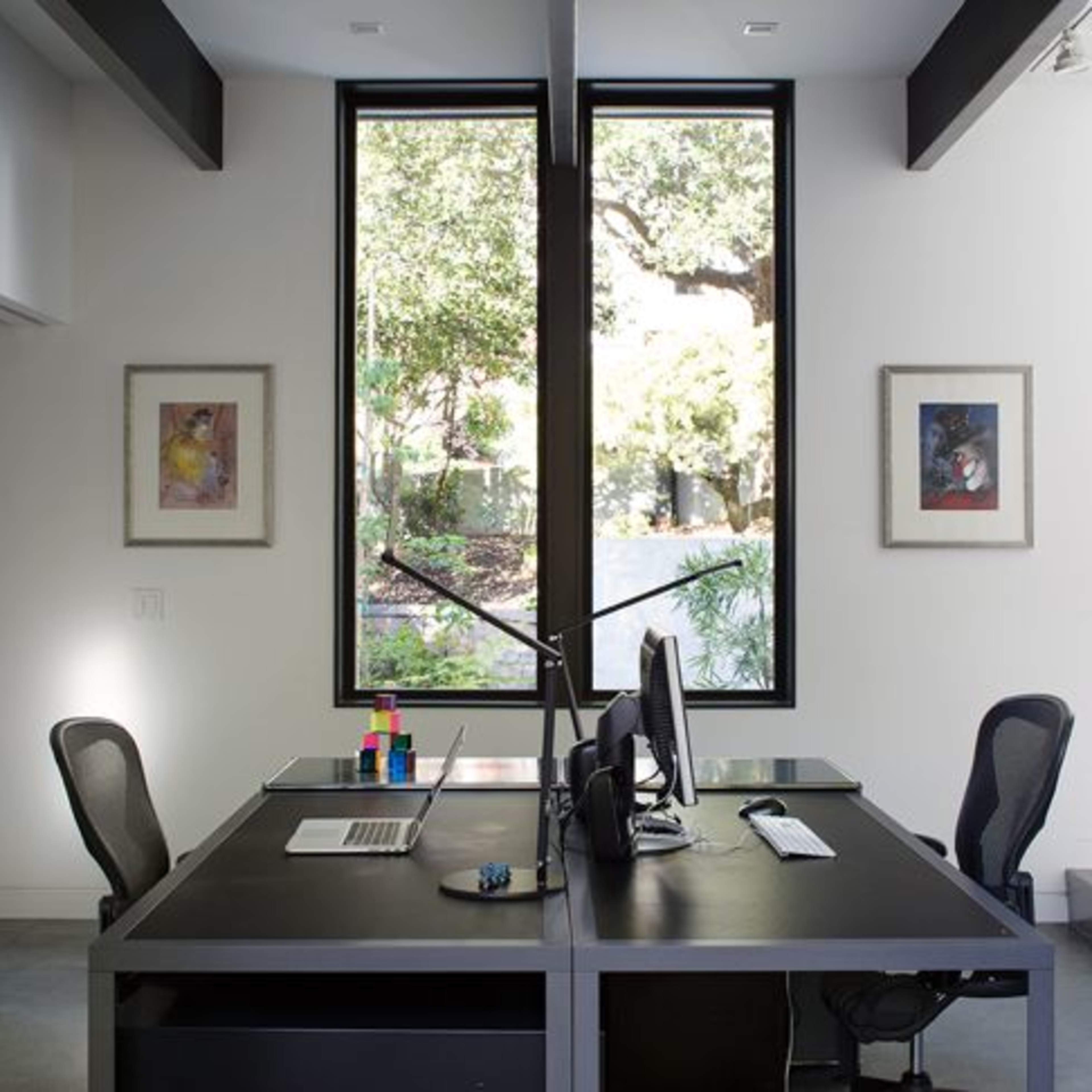 A modern office space features two desks facing large windows overlooking a garden.
