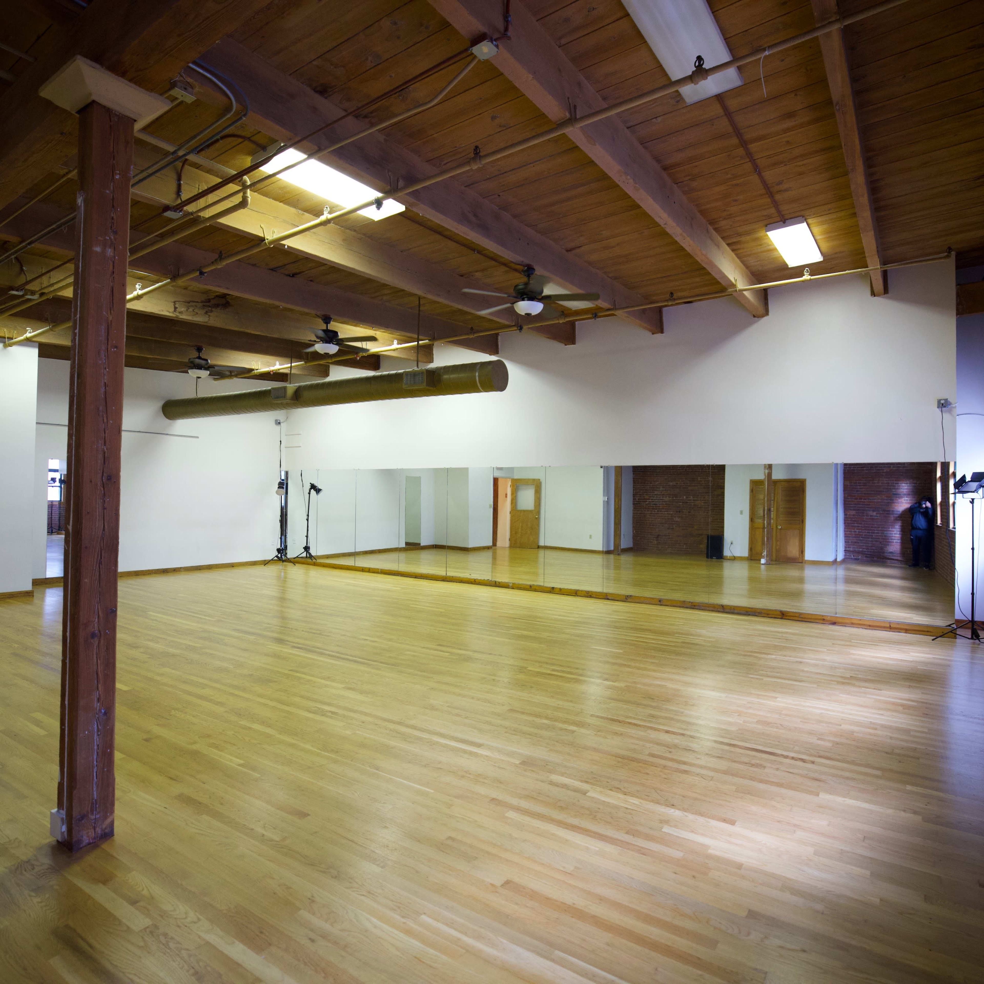 An empty dance studio with wooden floors, exposed beams, and large mirrors on one wall.