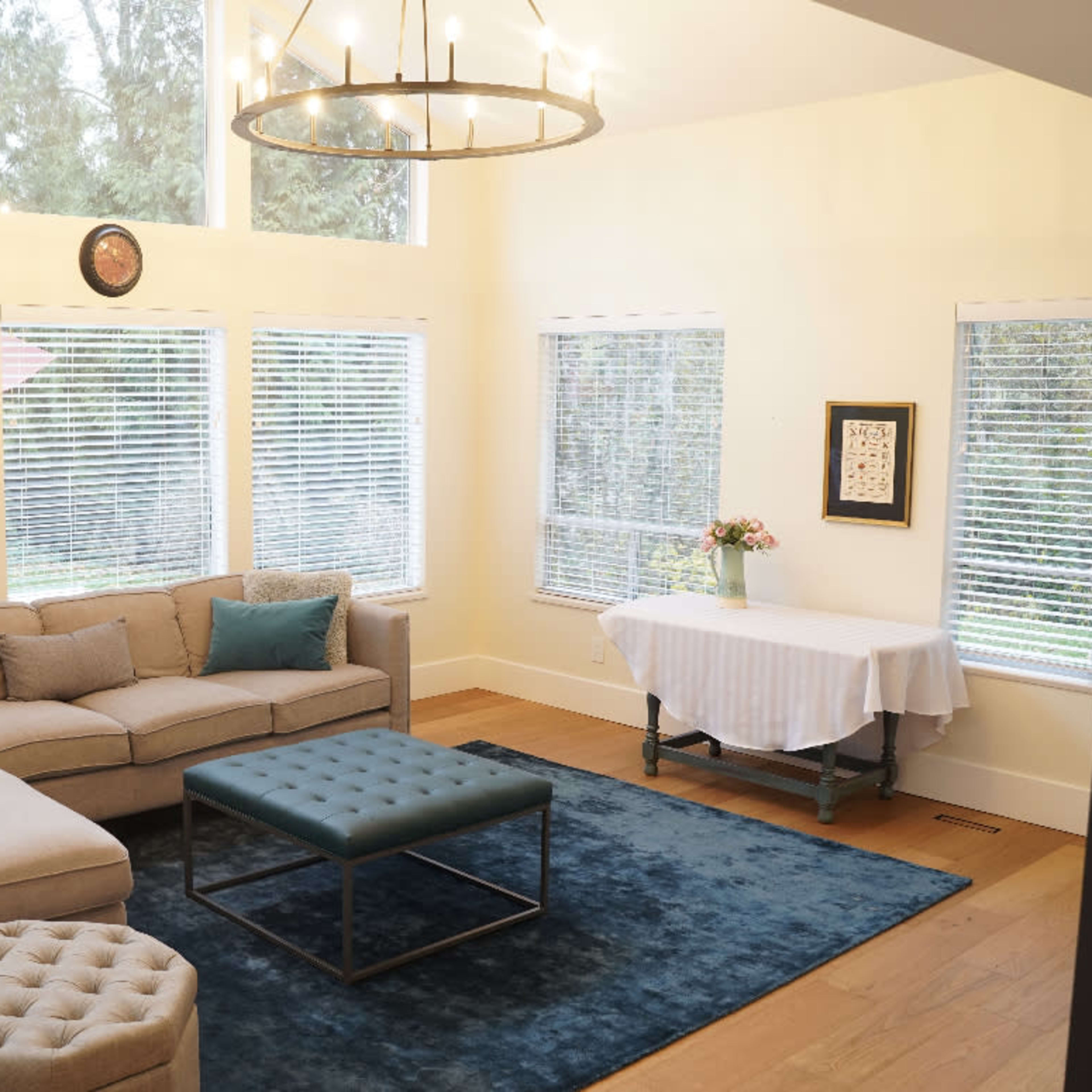 A modern living room features large windows, a gray sectional sofa, a blue rug, and a small dining table with flowers.